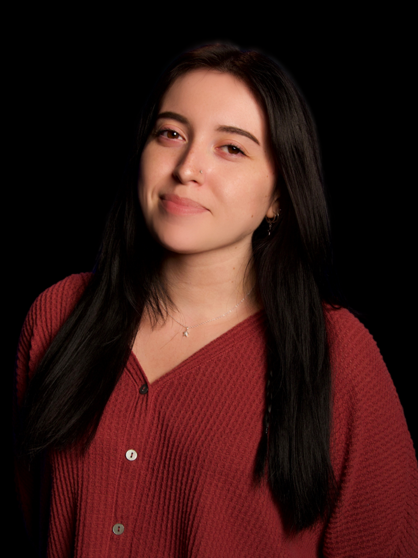 A young woman with long black hair, wearing a red button-up sweater, a delicate necklace, and earrings, smiling subtly against a black background.