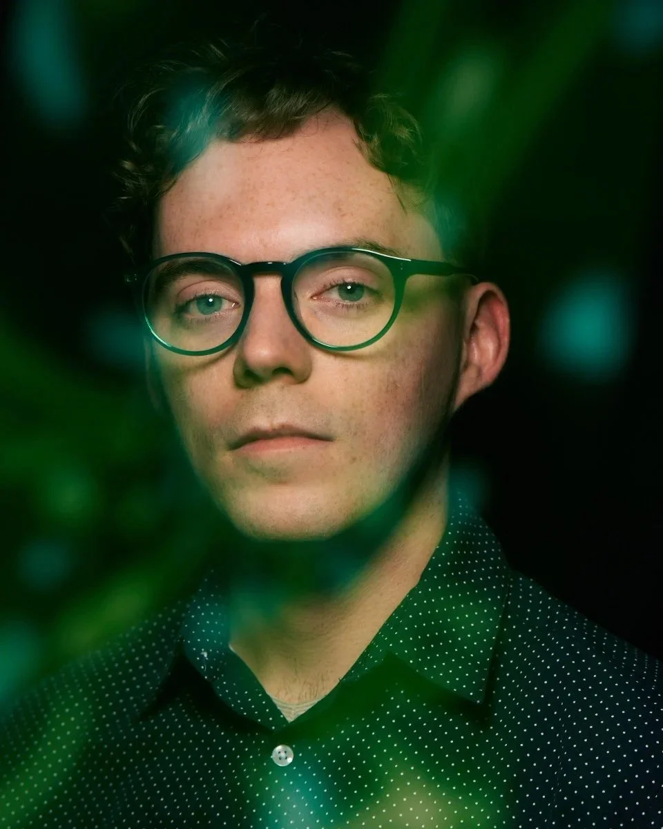 A young man with glasses and short curly hair, wearing a dark green shirt with white polka dots, looking directly at the camera with green leaves partially framing the foreground.