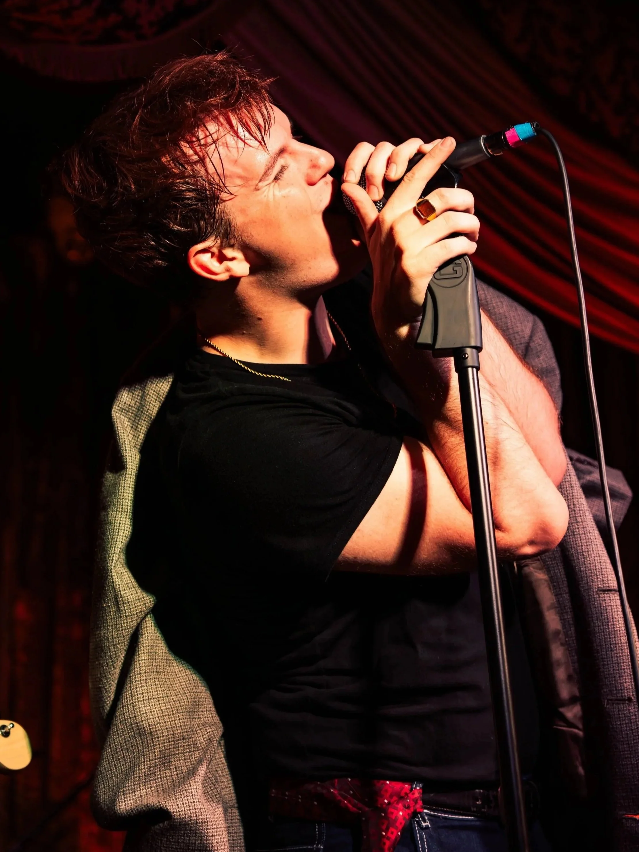 A man singing into a microphone during a performance, with a red curtain background.
