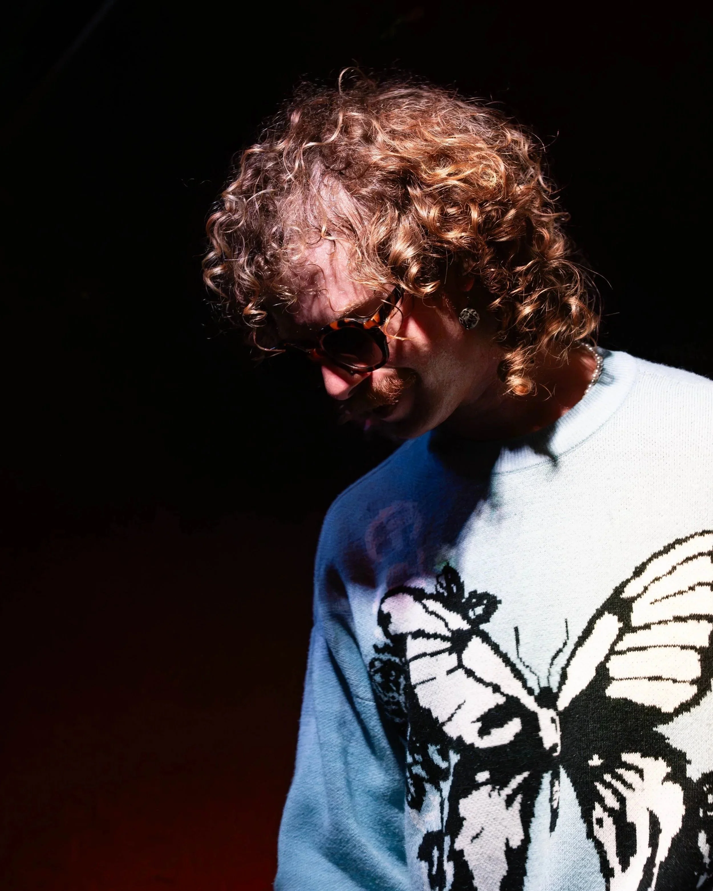 A person with curly red hair wearing sunglasses, a pearl necklace, and a white sweater with a black graphic design, looking downward against a dark background.