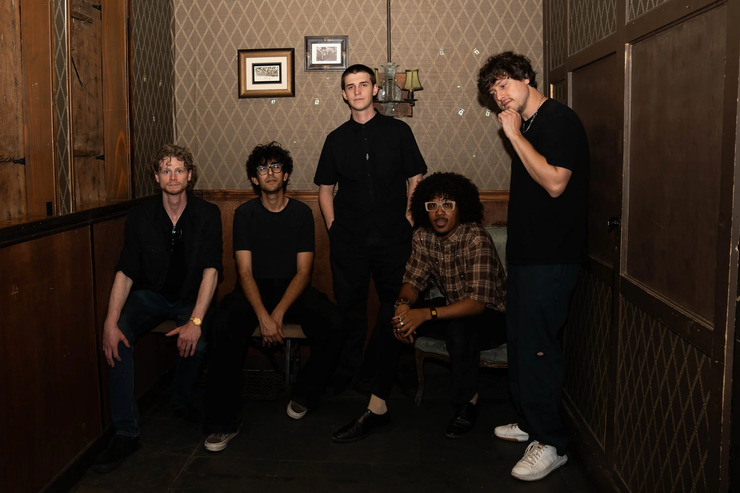 A group of five young adults, four men and one woman, posing in a small, vintage-style room with patterned wallpaper, wooden accents, and framed pictures. They are dressed in casual dark clothing.