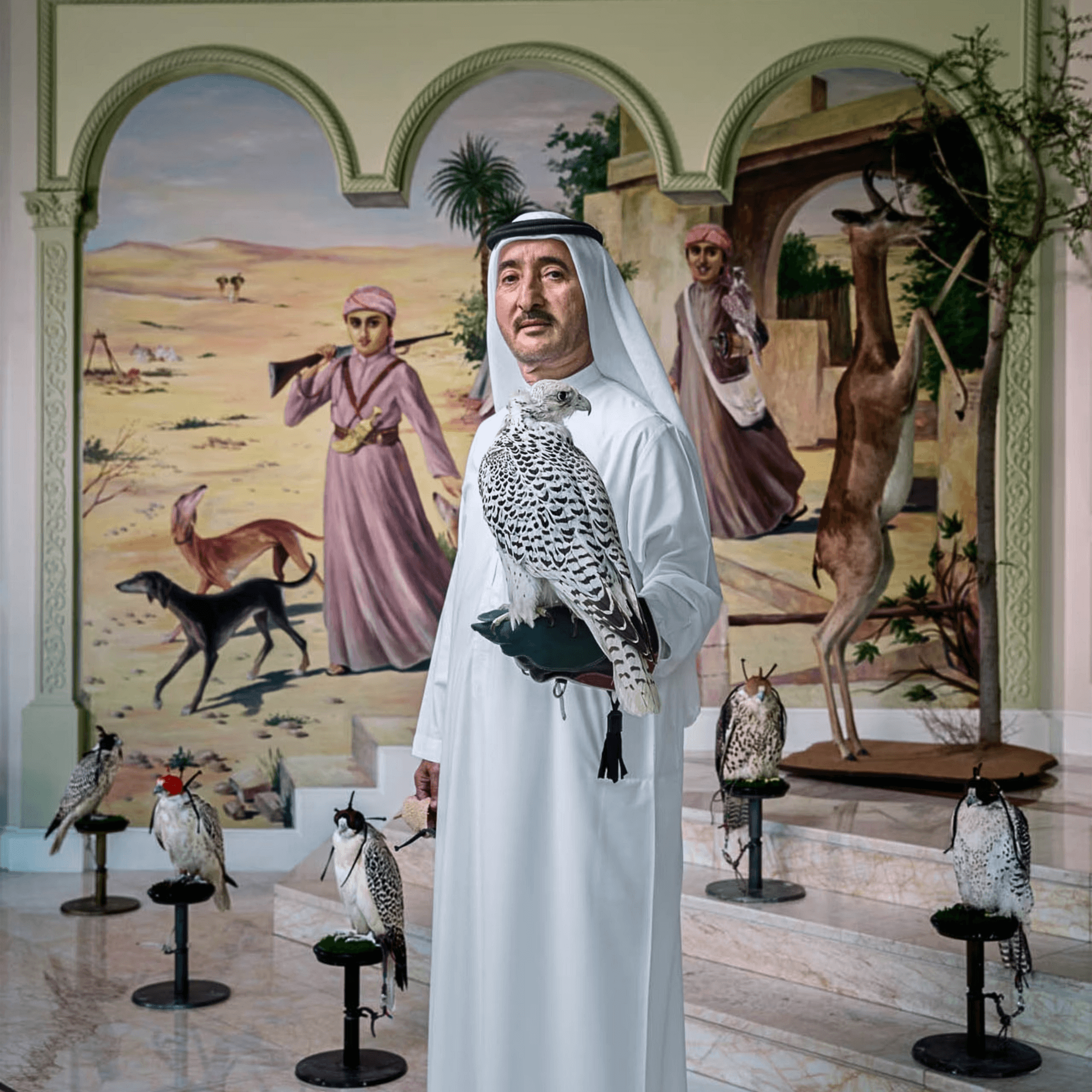 Indoor display of taxidermy animals including a falcon perched on a person’s gloved arm, surrounded by other mounted animals including a deer, fox, and rabbits, with a painted desert landscape background and arches.
