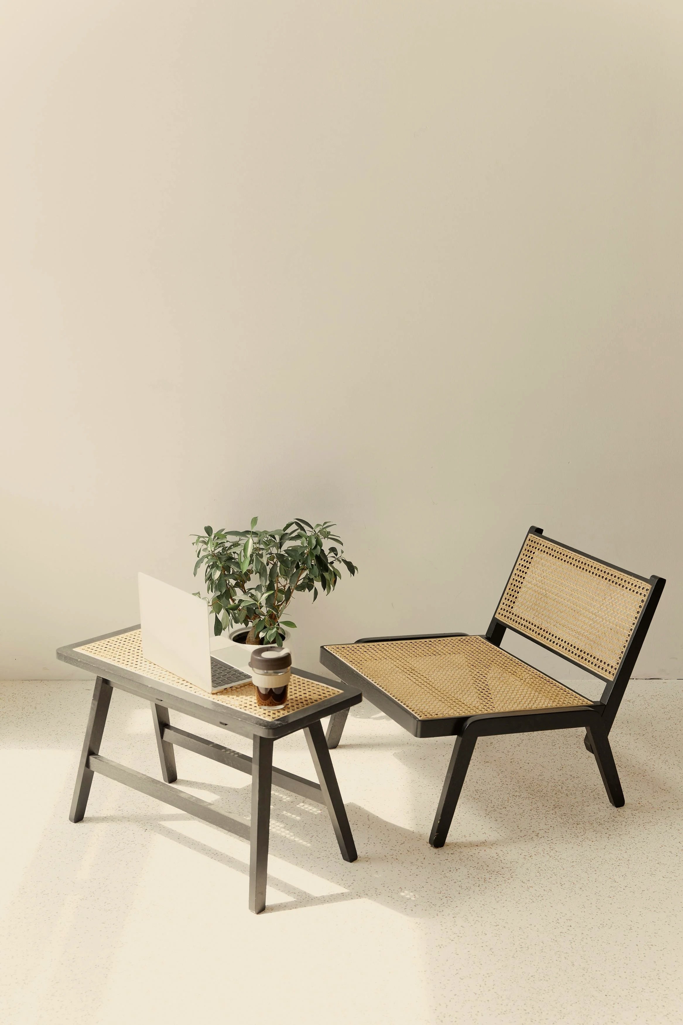 An office space with a small table holding a laptop, a coffee cup, and a potted plant, with a chair next to it, against a plain light-colored wall.
