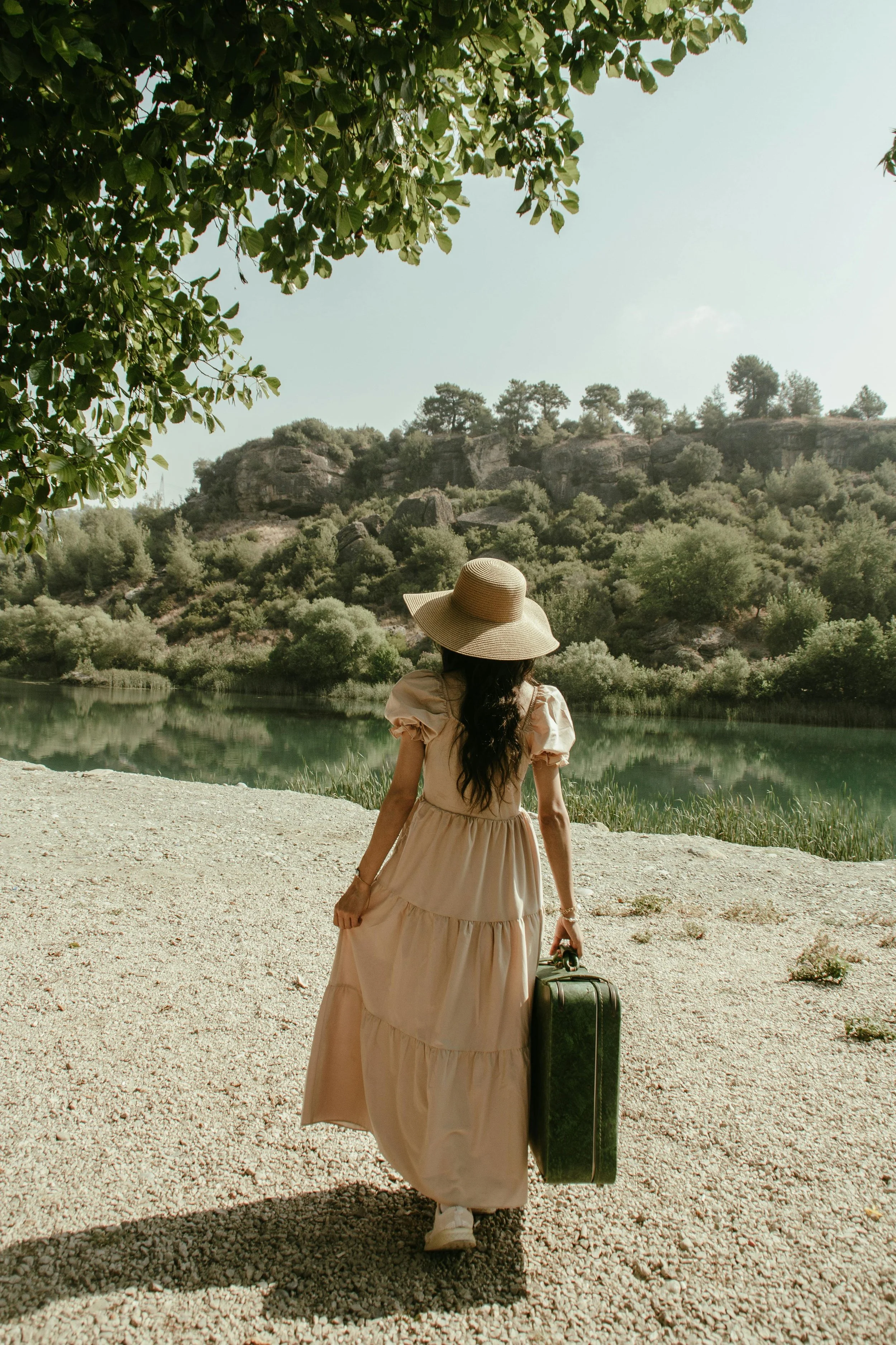 Woman wearing a beige dress, wide-brimmed hat, white sneakers, and carrying a vintage suitcase walking along a gravel path beside a lake with forested hills in the background.