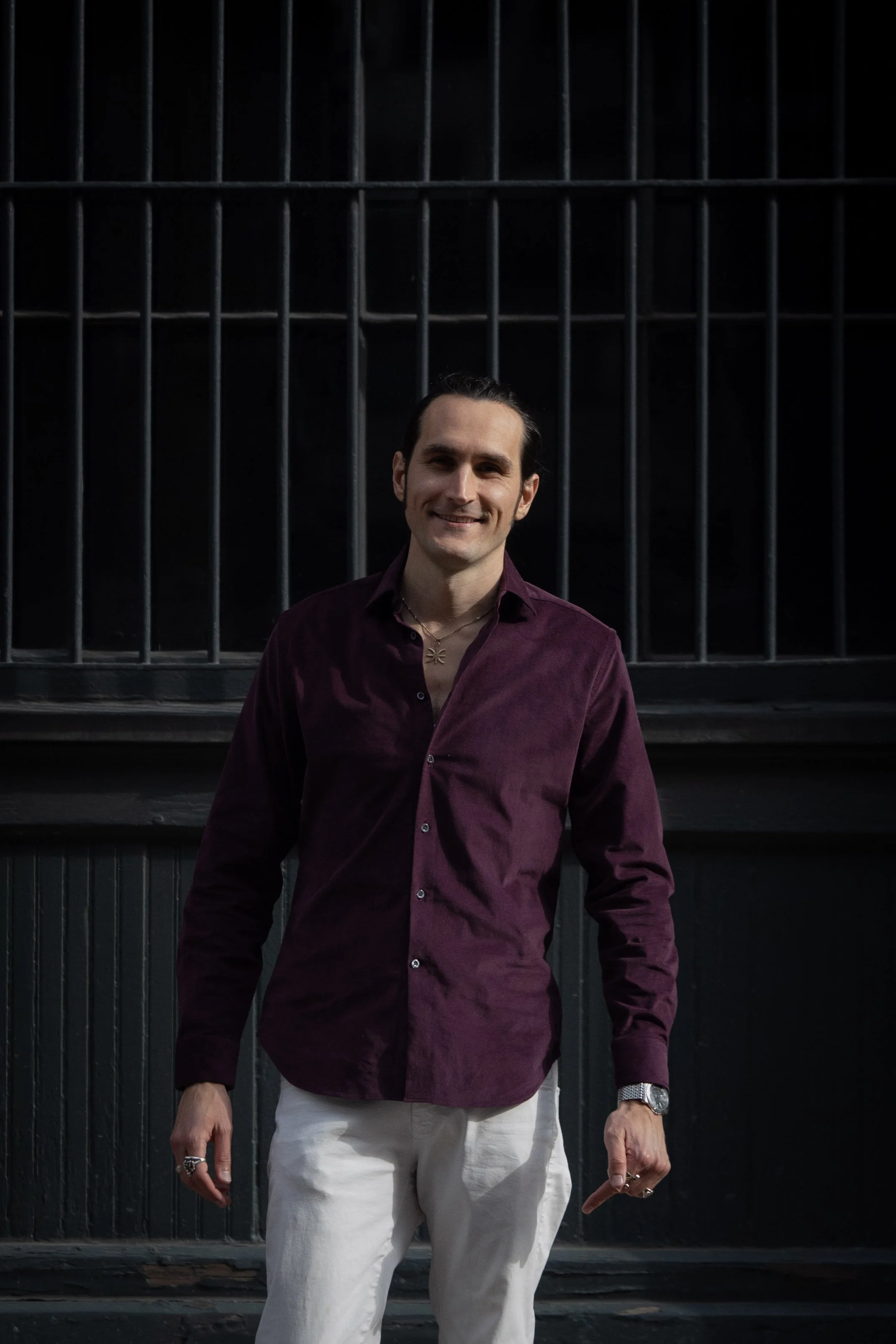A man with dark hair and light skin is smiling and standing in front of a dark iron gate.