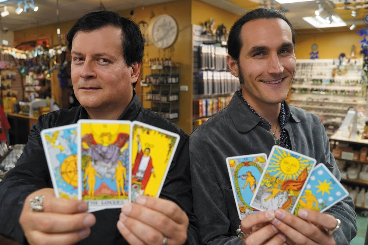 Two men holding tarot cards inside a store, with shelves and a yellow wall in the background.