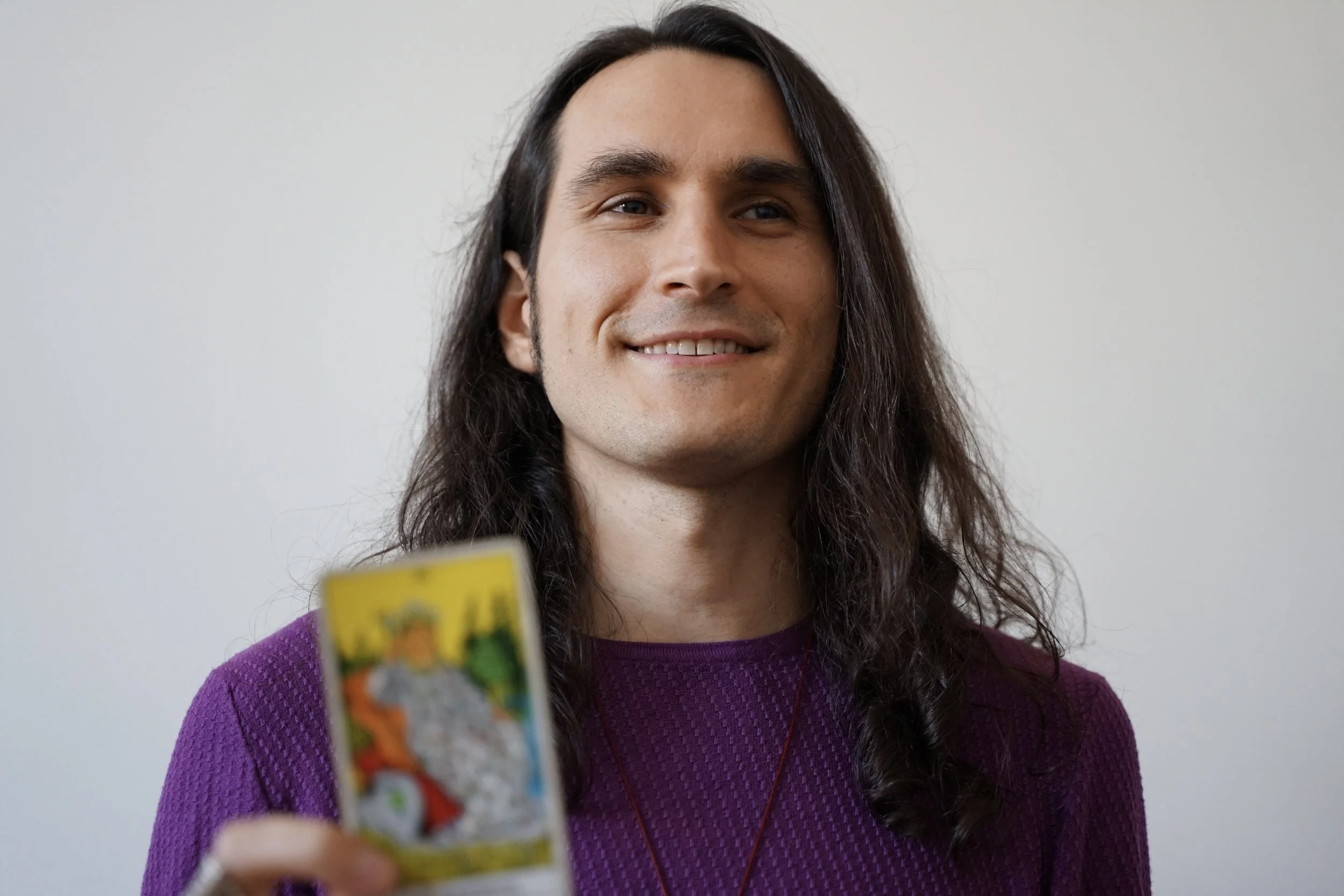 A young man with long dark hair, wearing a purple sweater, smiling, with a plain white background. He is holding a box of colorful snacks close to the camera.