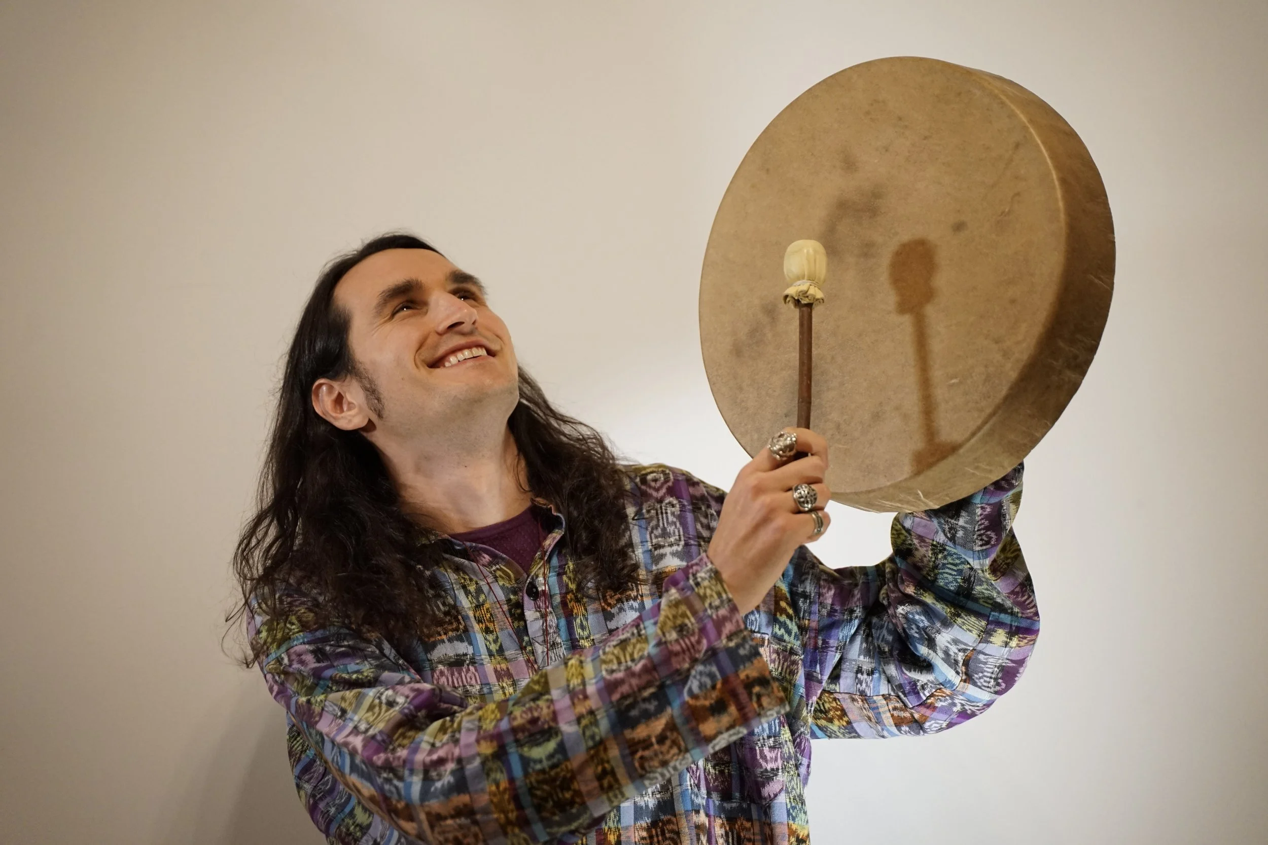 A man with long dark hair, smiling and looking up, holding a large gong with a mallet in front of a plain wall.