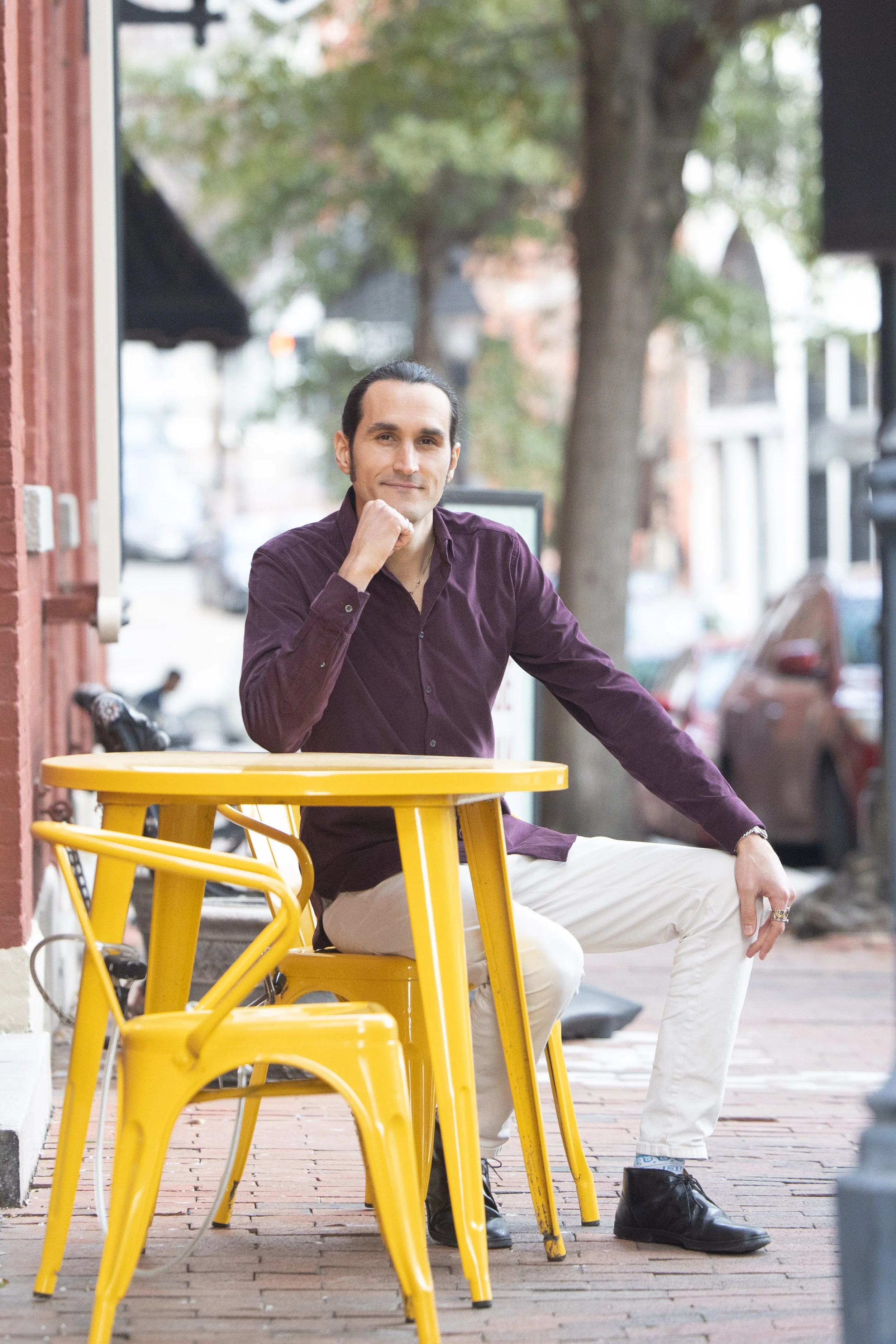 A man with dark hair pulled back, wearing a dark purple shirt and light-colored pants, sitting on a yellow stool outside a café, leaning his chin on his hand and looking at the camera. There are yellow tables and chairs, a brick sidewalk, and trees in the background.