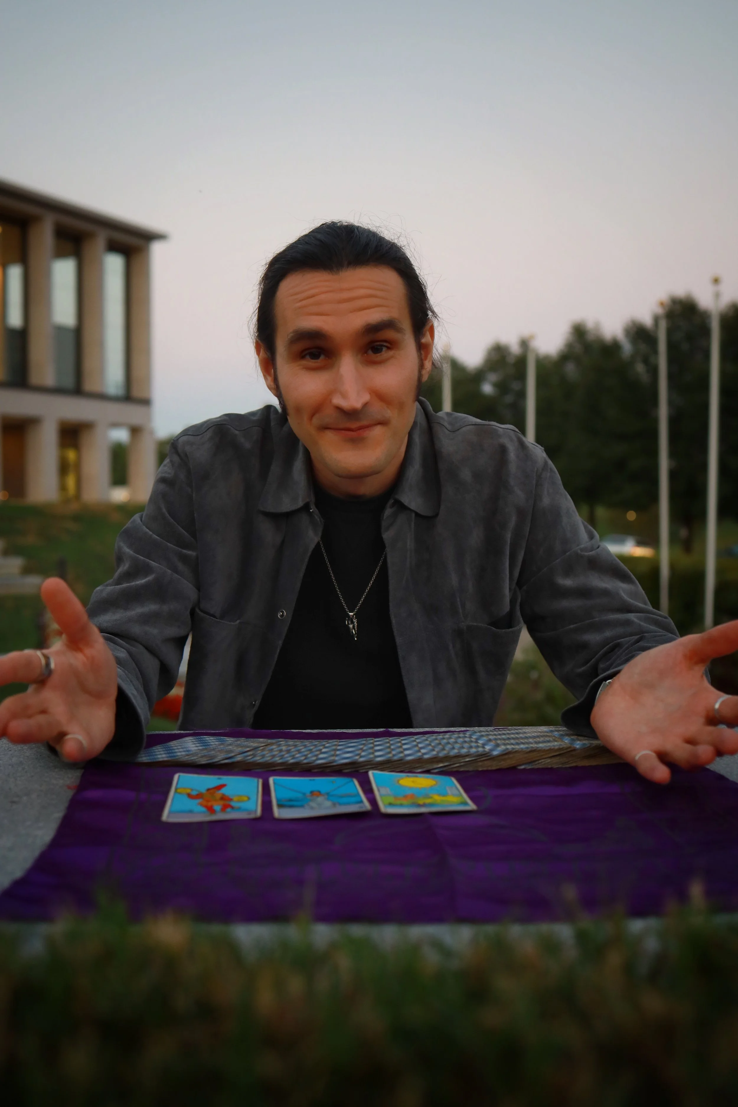 A man with dark hair and a slight smile, wearing a dark shirt and necklace, leaning forward with his hands open towards the camera, sitting outdoors in front of a table with tarot cards, with a modern building and flagpoles in the background during dusk.