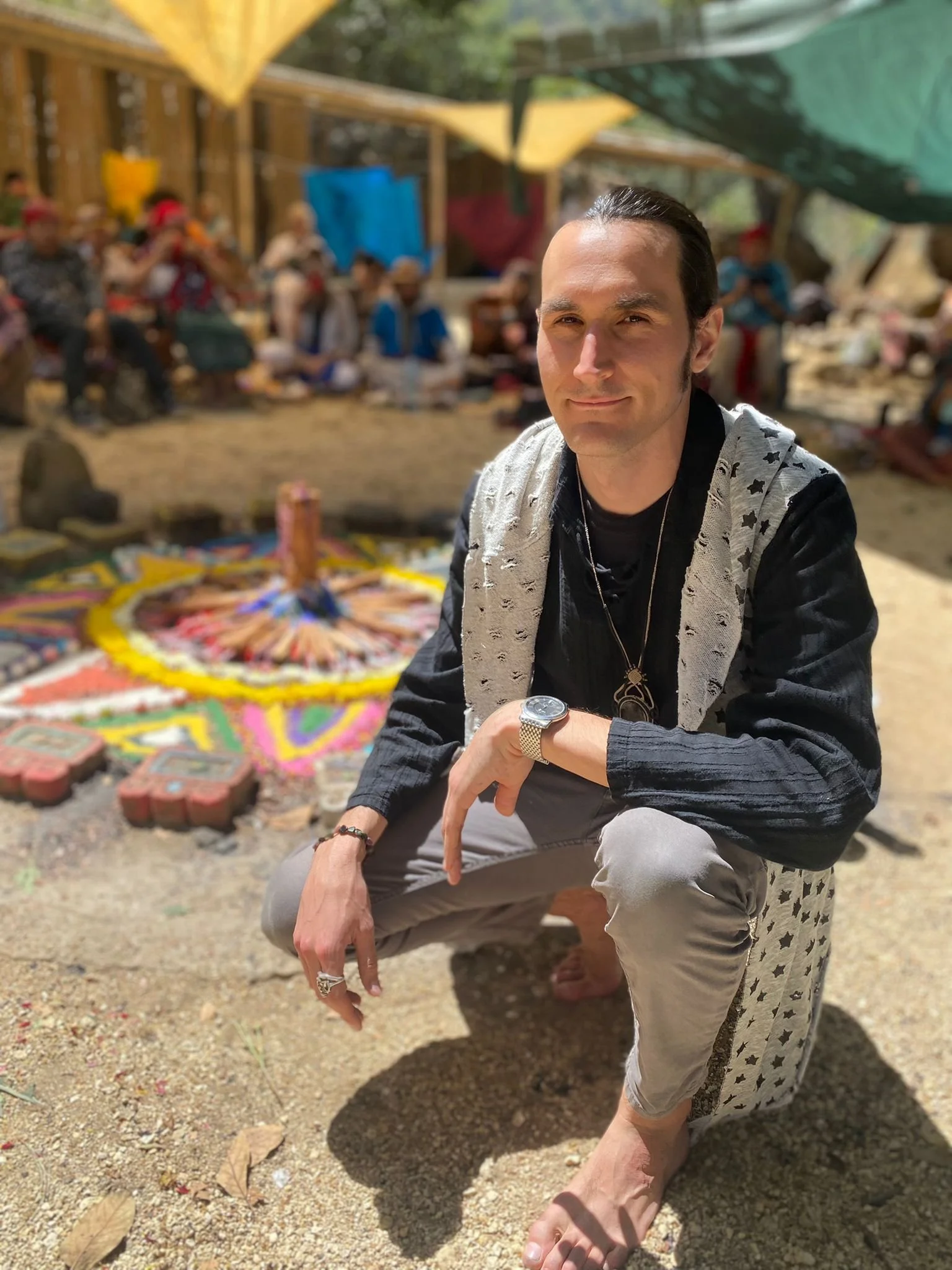 A man with slicked-back hair, wearing a black shirt and a jacket with star patterns, is sitting outdoors on a dirt ground, with a large circle of decorated items on the ground behind him. There are people in the background under a shaded area made of colorful fabric, and they appear to be participating in a gathering or event.