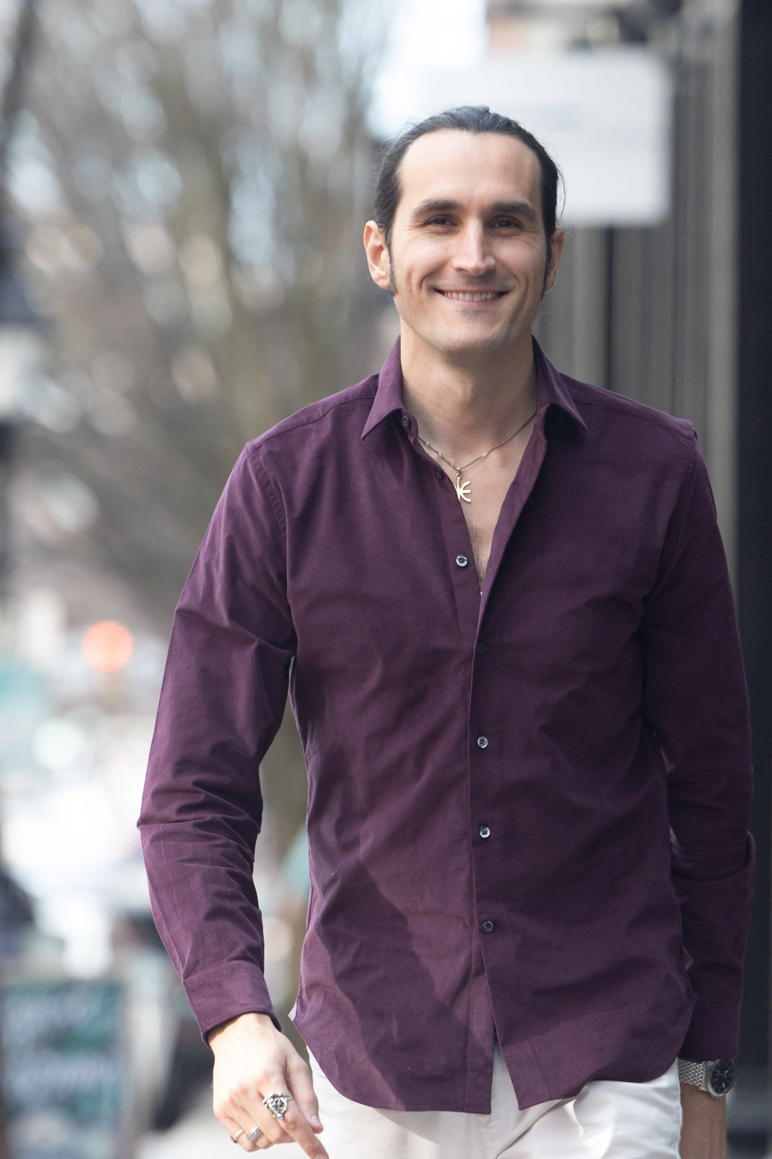 A man with long dark hair, wearing a purple button-up shirt, beige pants, and a necklace with a cross pendant, walking outdoors on a city street.