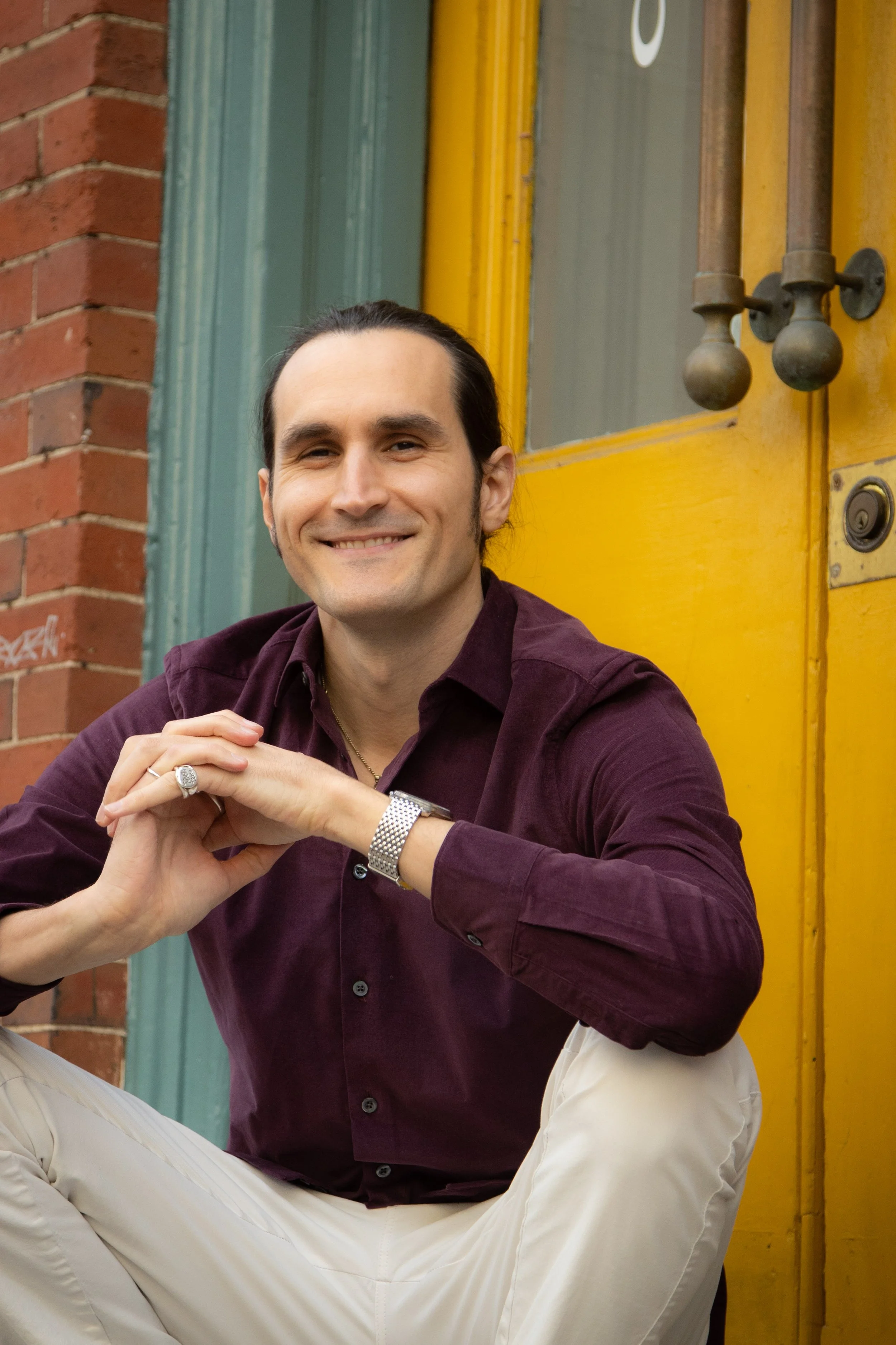 A smiling man with dark hair, dressed in a maroon shirt, sits with his hand resting on his knee outside a building with a yellow door and a brick wall.