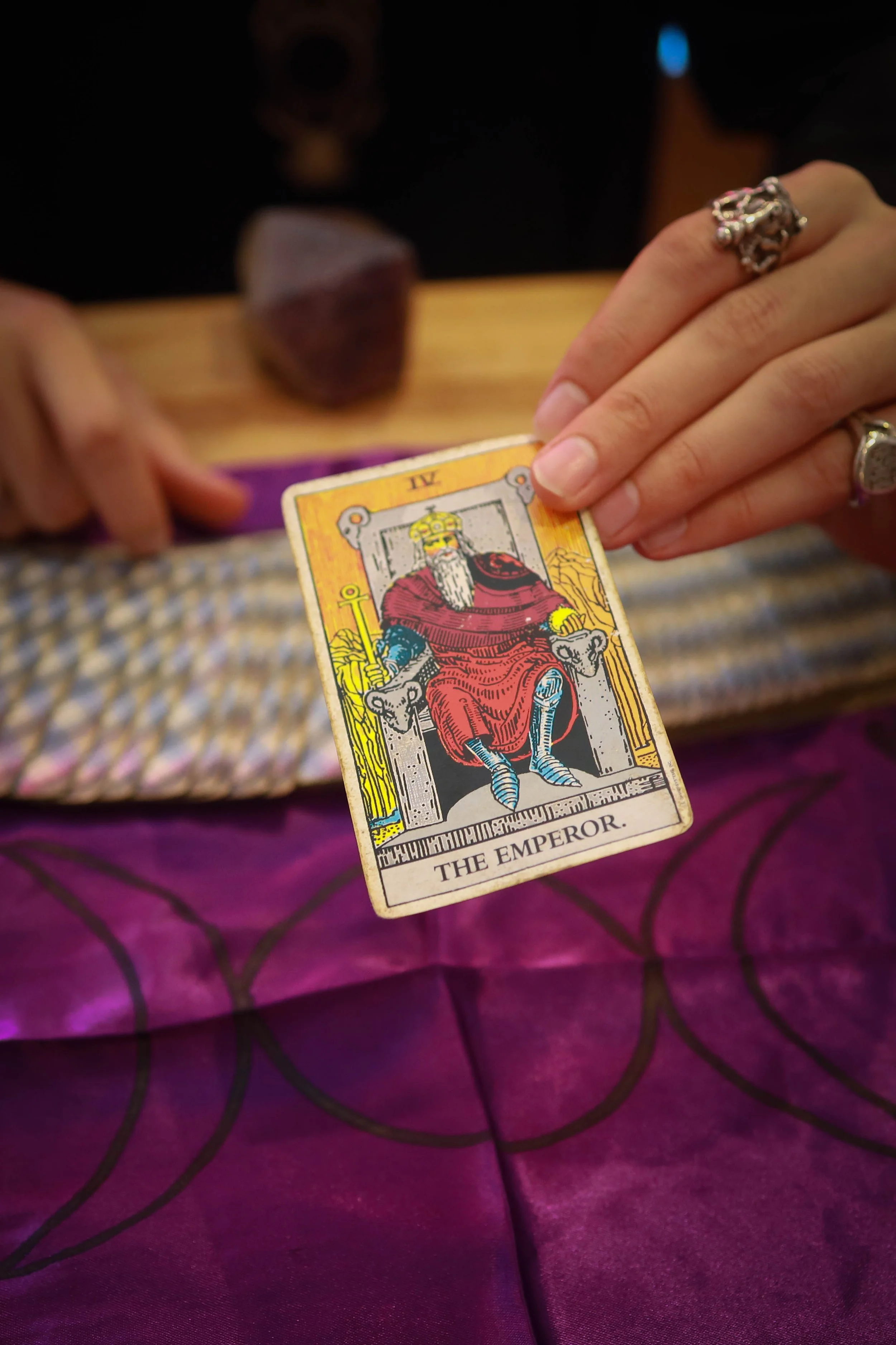 Close-up of a tarot card titled 'The Emperor' held by a person's hand, with rings on fingers, on a purple cloth surface.