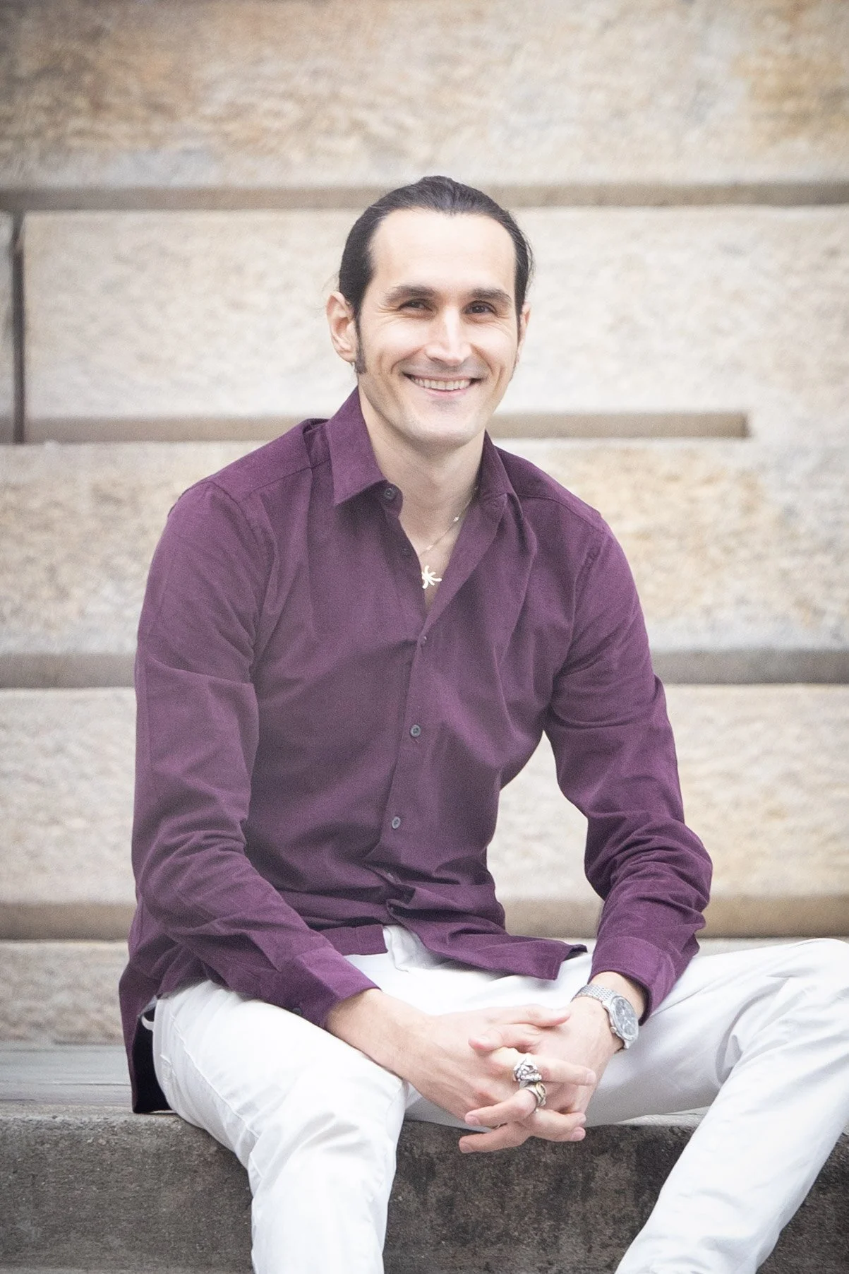 A man sitting on concrete stairs outdoors, smiling, wearing a purple button-up shirt and white pants.