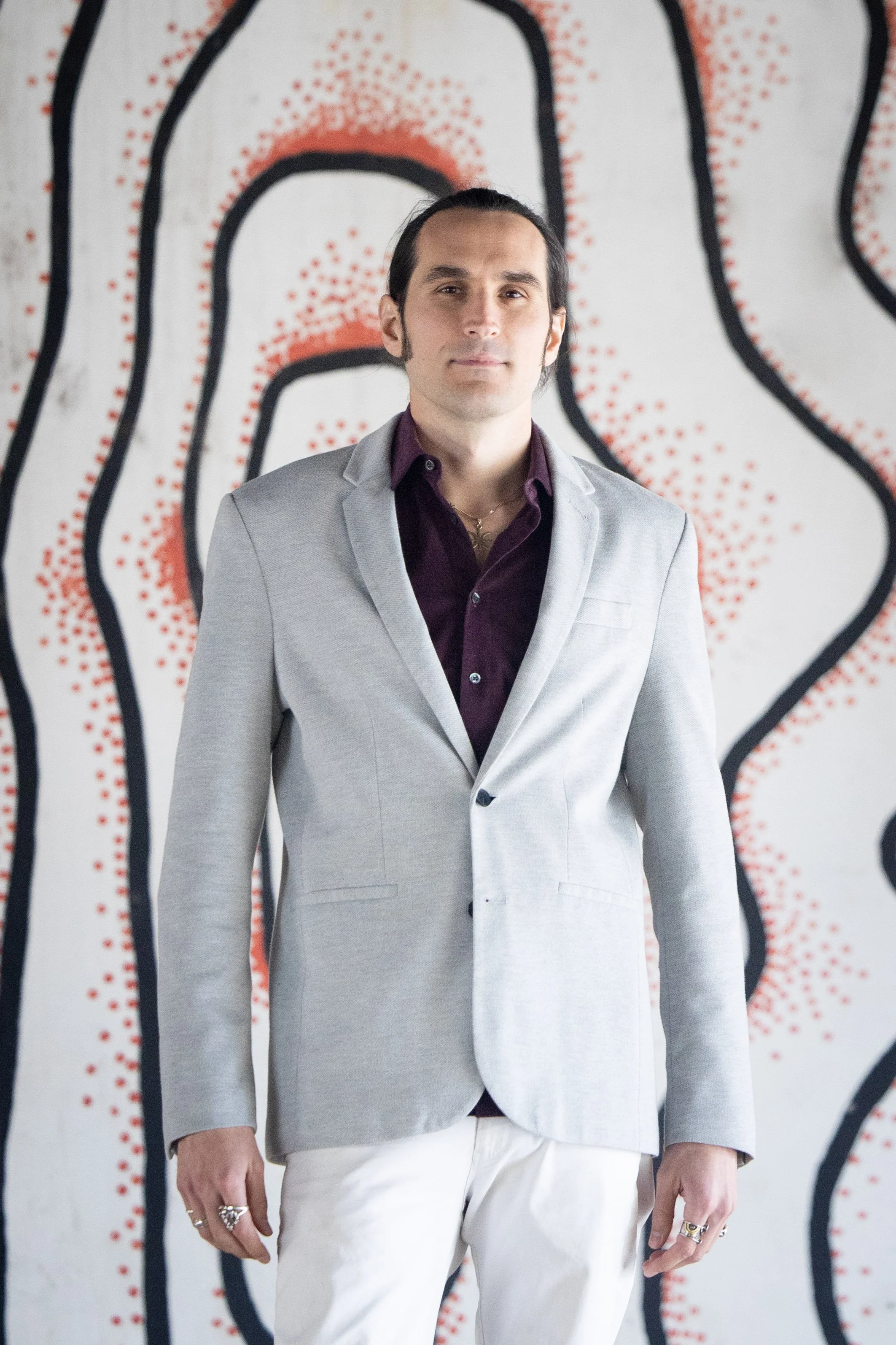 A man in a light gray suit with a purple shirt standing in front of an abstract black, red, and white mural.
