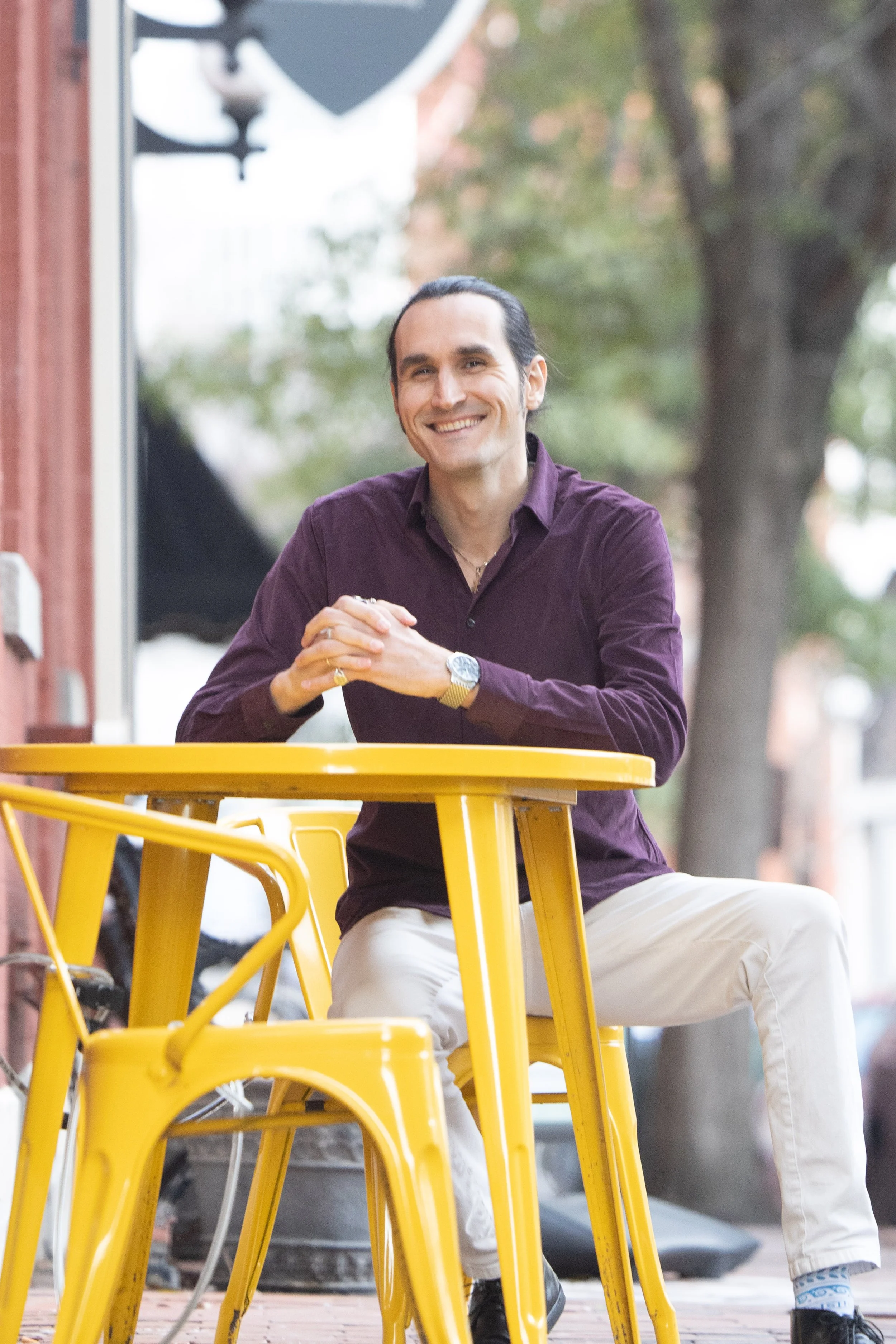 A man with dark hair and a broad smile sitting at a yellow outdoor table, wearing a dark purple shirt and light-colored pants. He has a watch and a ring on his left hand. In the background, there are trees and outdoor elements.