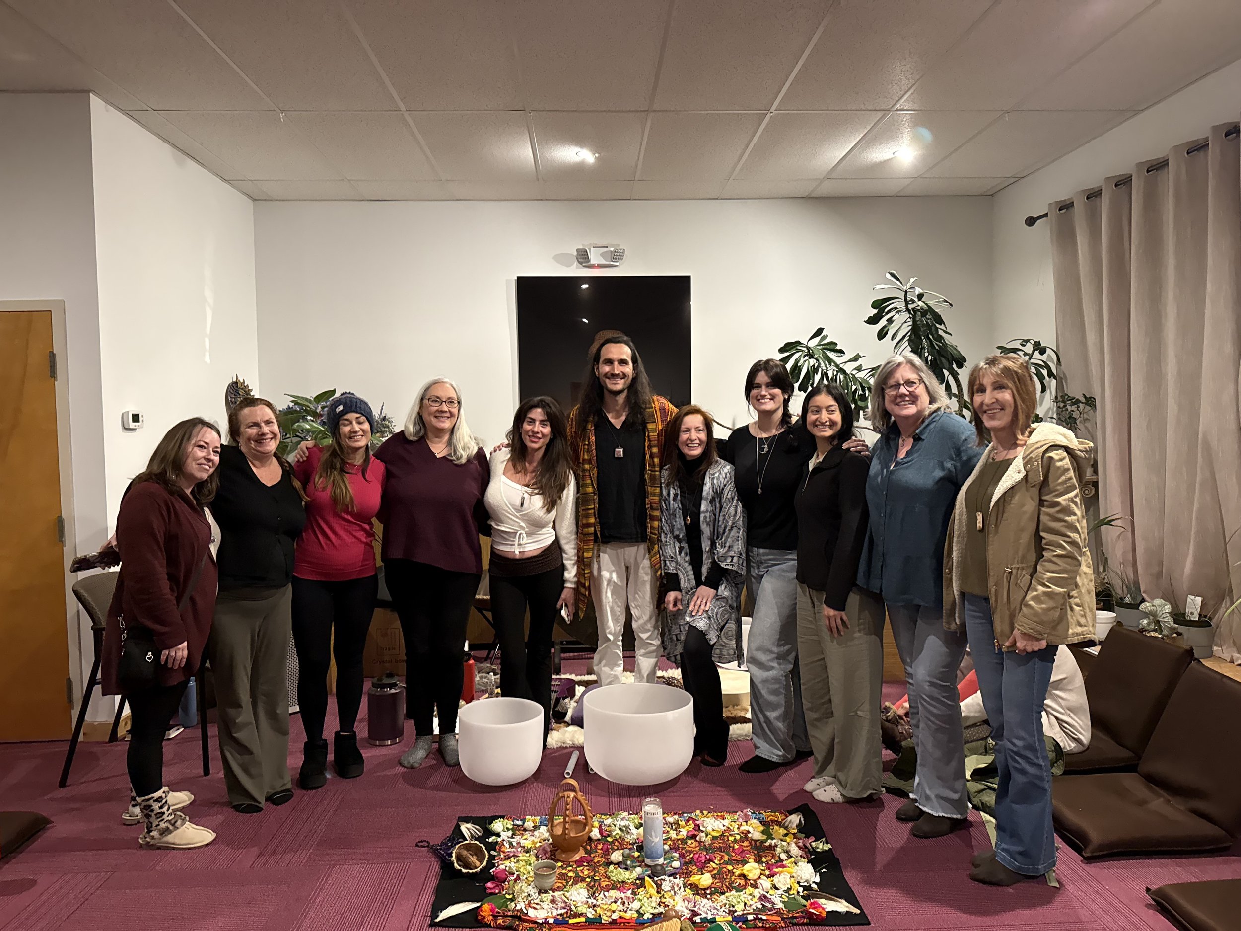 A group of people standing together inside a room, posing for a photo, with a decorated altar or display in front of them, surrounded by plants, with a television and beige curtains in the background.