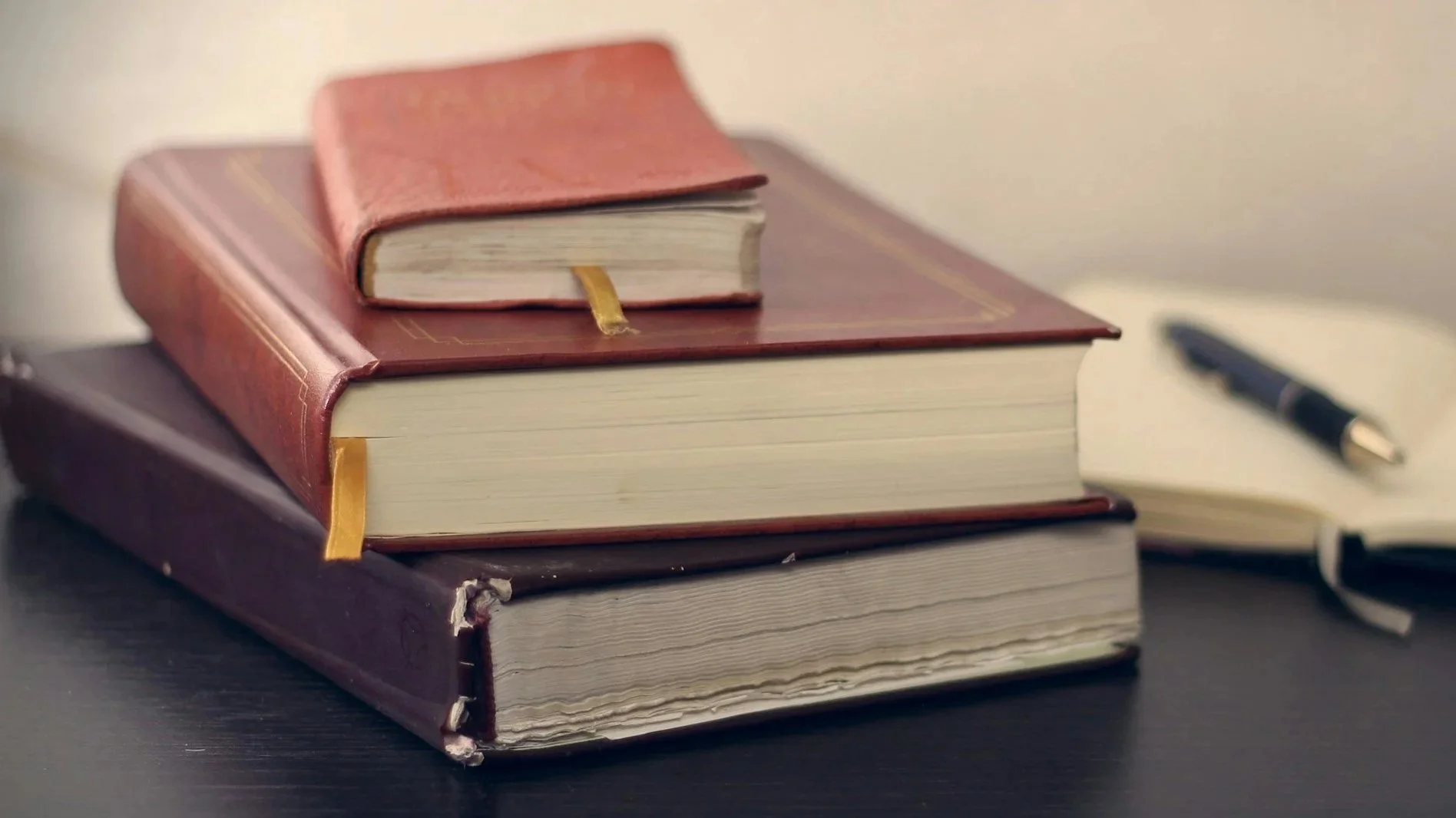 Stack of three hardcover books with a small red leather book on top, a black pen to the right, and a notebook in the background on a dark surface.