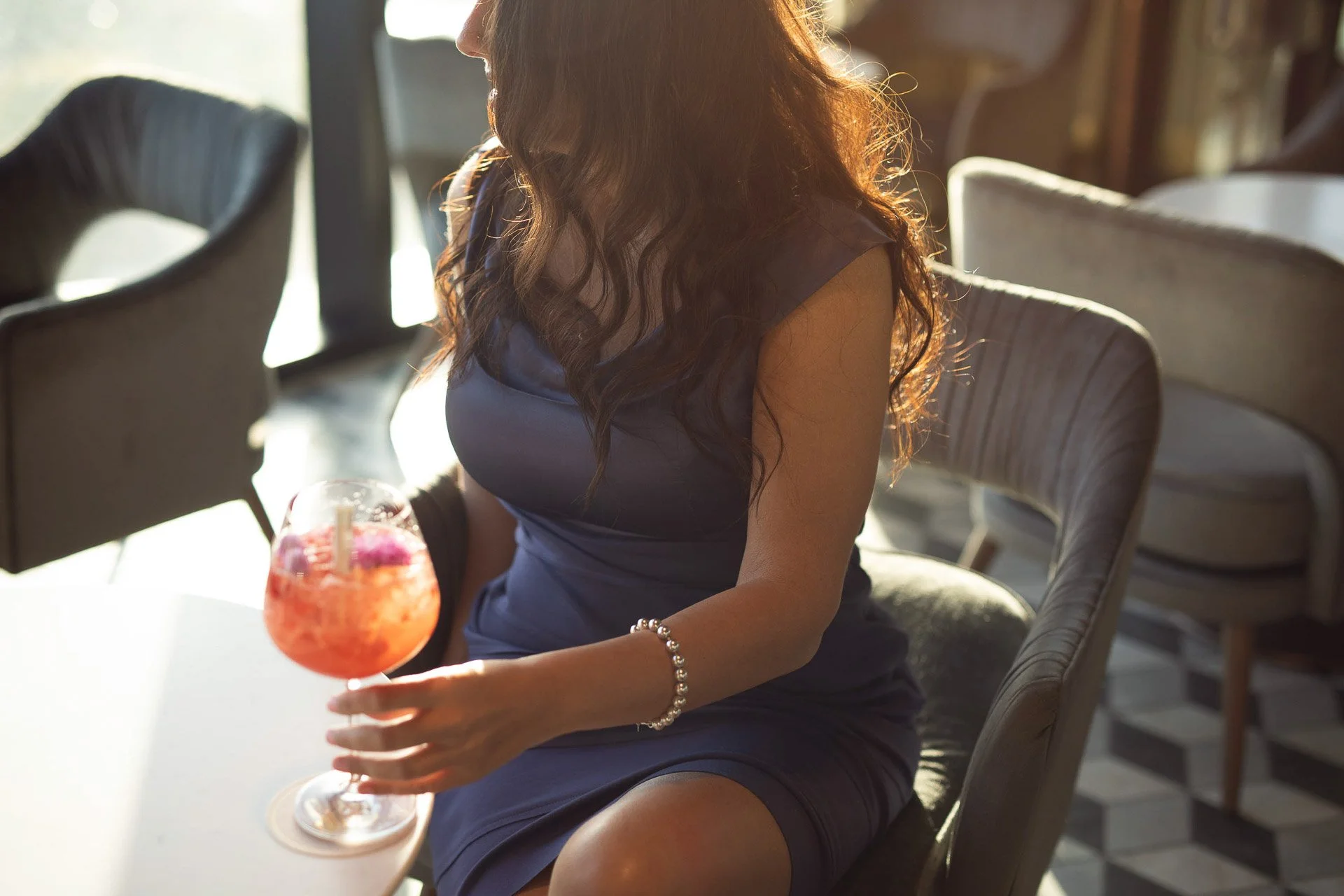 Carla Rinaldi with long, wavy hair, wearing a dark dress, holding a colorful cocktail drink in a glass, sitting in a well-lit café or restaurant.