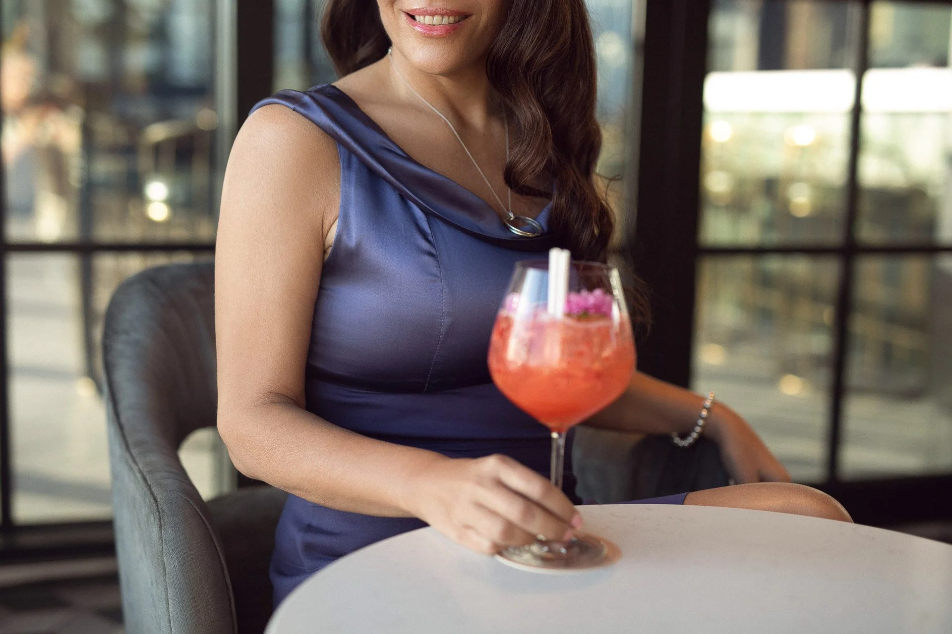 Carla Rinaldi in a blue satin dress holding a pink cocktail garnished with an edible flower in a restaurant with large windows and natural light.
