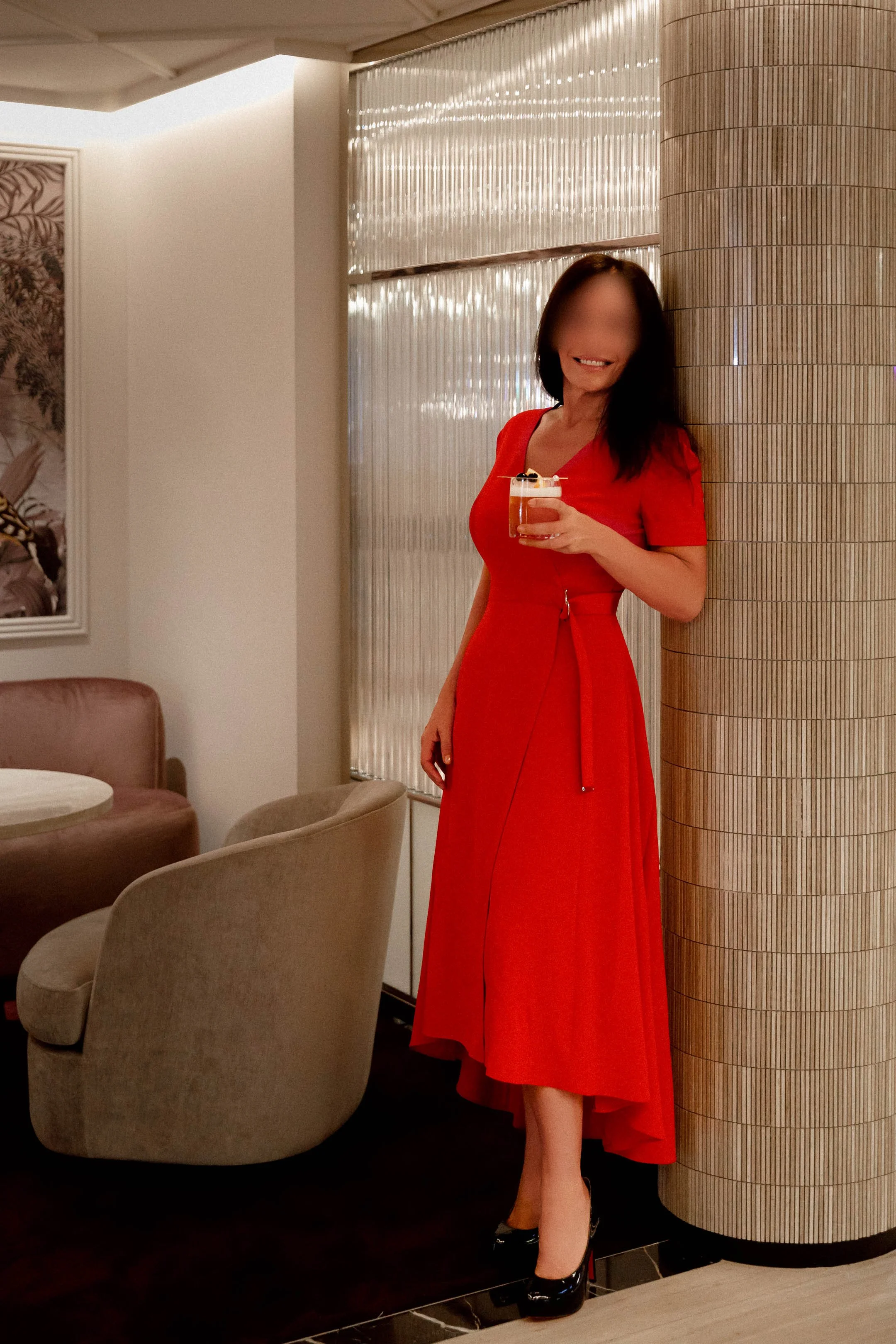 Carla Rinaldi in a long red dress and high heels is holding a drink while standing next to a curved wooden pillar inside a restaurant or lounge.