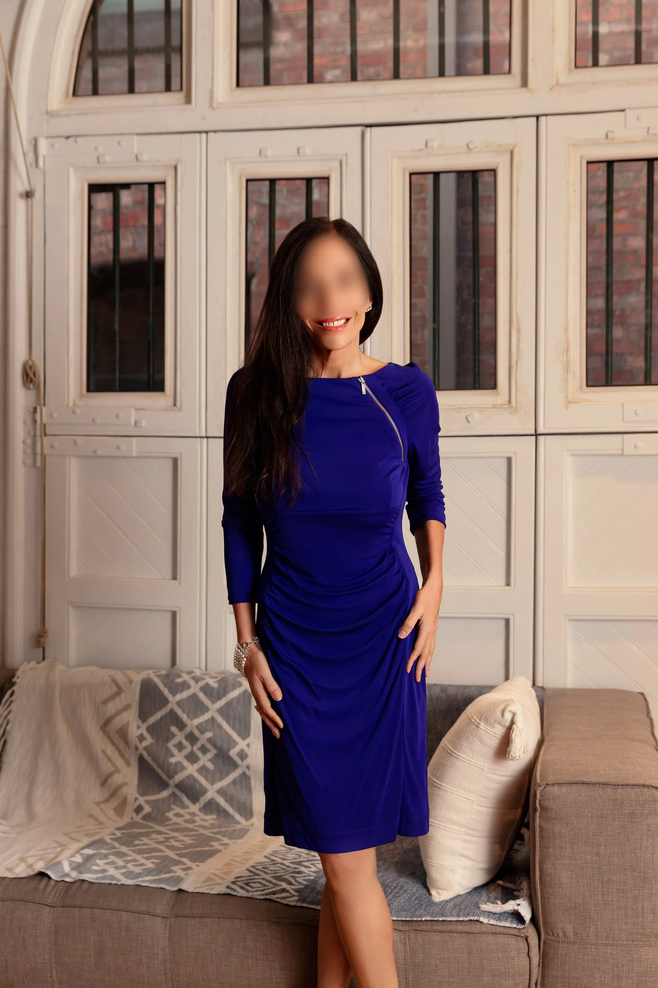 Carla Rinaldi in a royal blue dress standing in a cozy living room with a sofa, pillows, and a decorative blanket, in front of a large window with wooden panels.