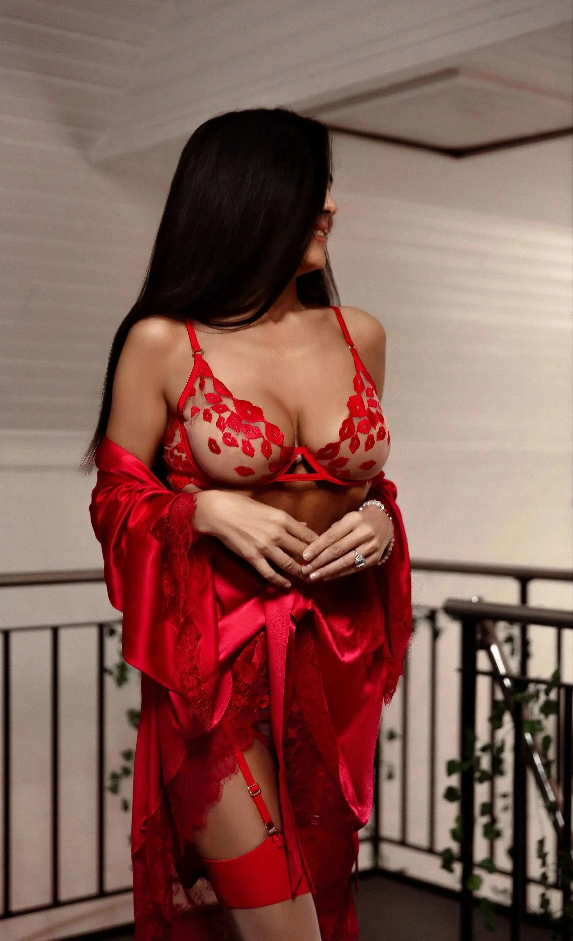 Carla Rinaldi in red lingerie and robe, standing on a balcony, smiling and looking to her left.