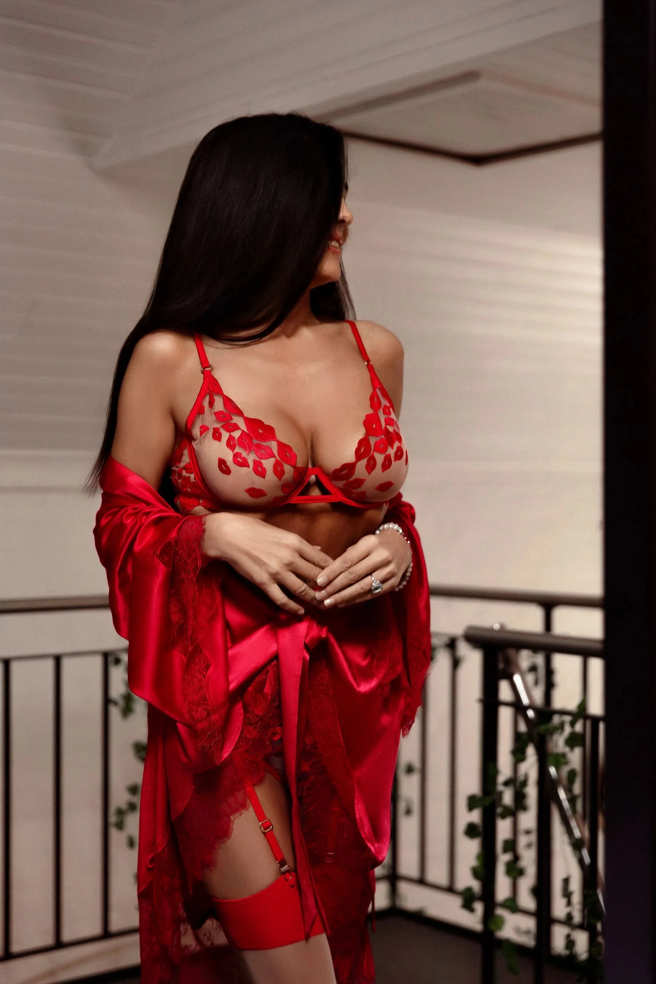 Carla Rinaldi wearing red lingerie and a satin robe standing on a balcony, facing sideways, with long dark hair.