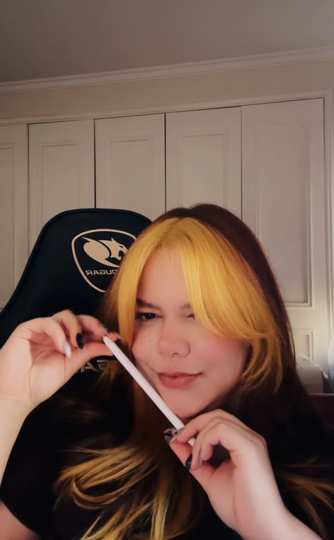 Mujer con cabello rubio y oscuro, sonriendo, sosteniendo un cotonete, en una habitación con puertas blancas y una silla Gaming.