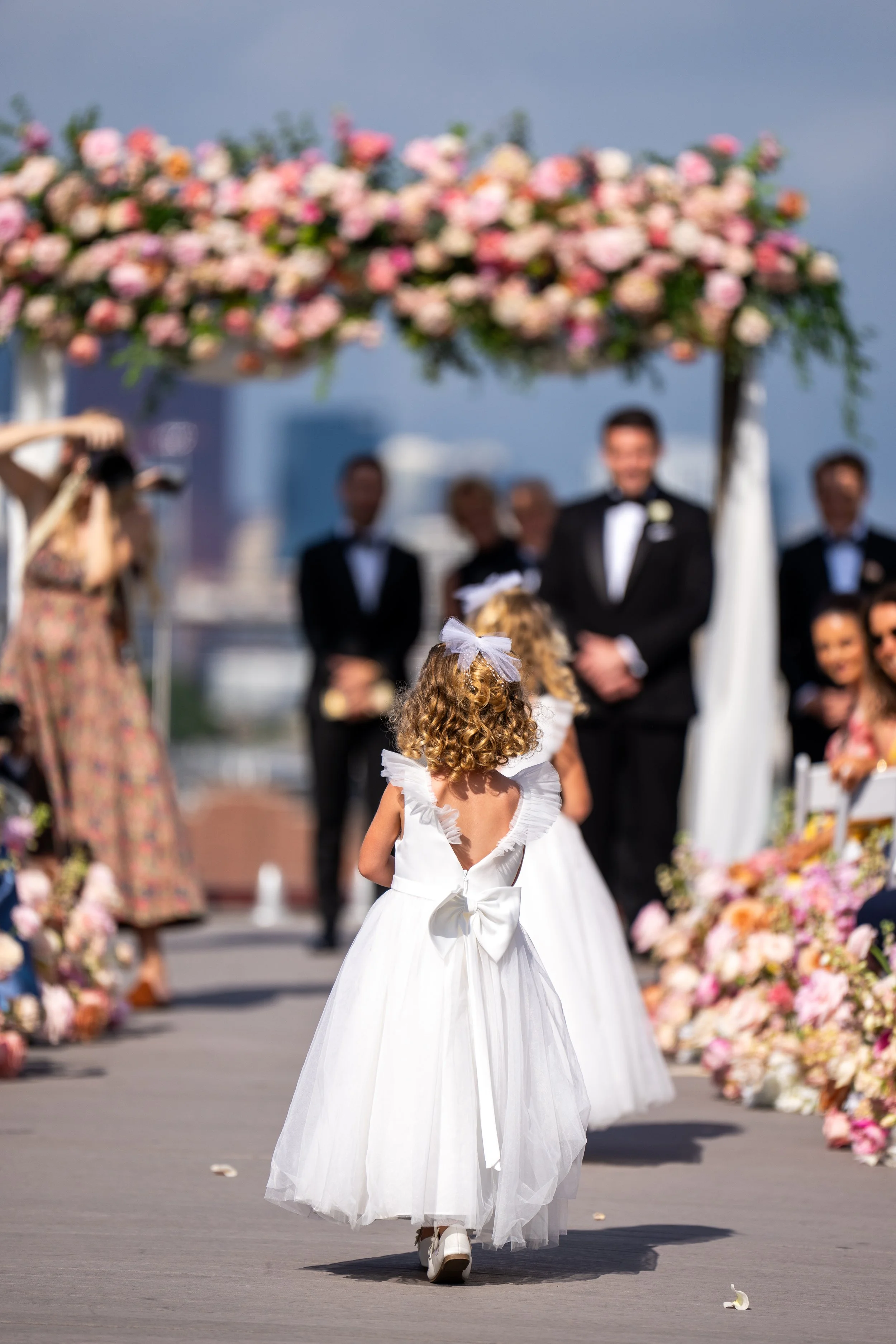 A young girl in a white dress with a bow at the back walking in front of a wedding ceremony setup with pink and peach flowers and men in tuxedos, under a floral arch.