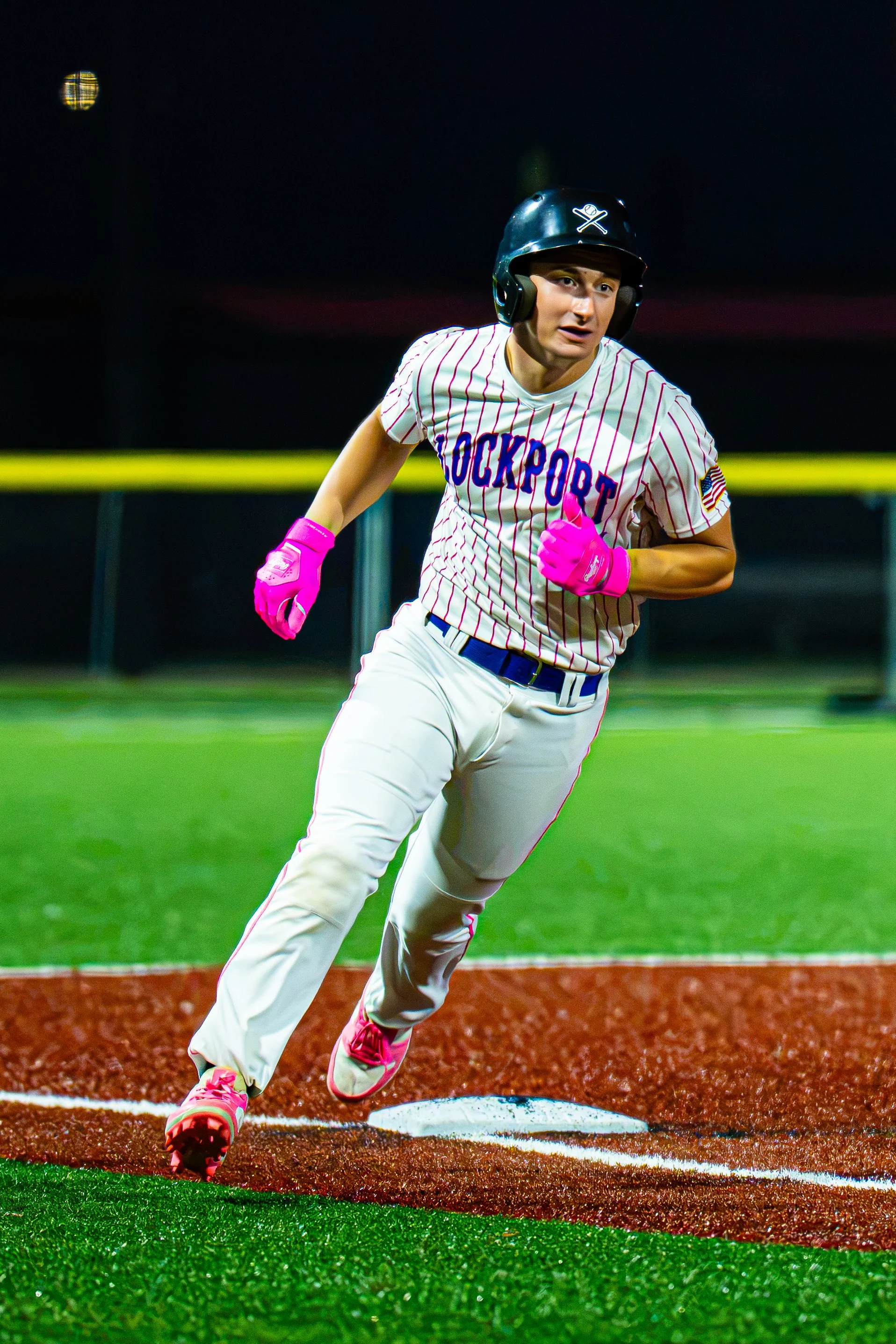 A baseball player wearing a white pinstriped uniform with 'ROCKPORT' written on it, pink gloves, pink shoes, and a black helmet, is running from third base to home plate during a game at night.