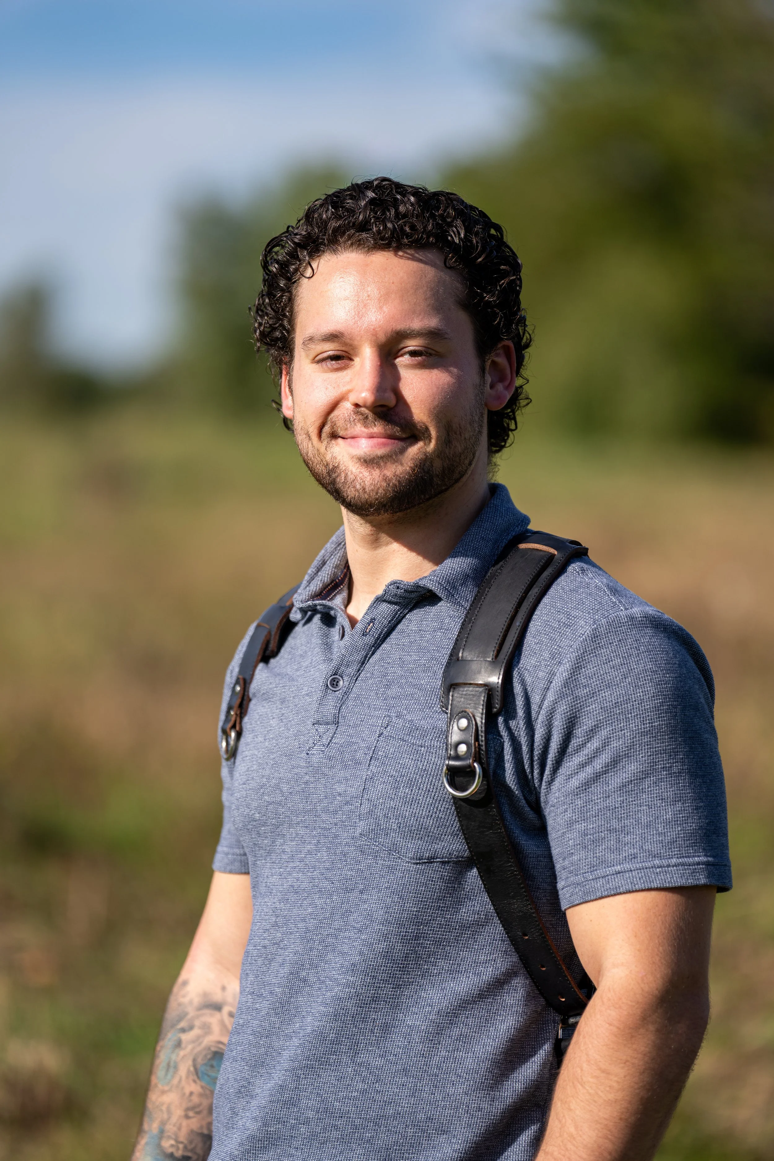 A young man with curly dark hair and facial hair, standing outdoors with a backpack, smiling and looking at the camera.