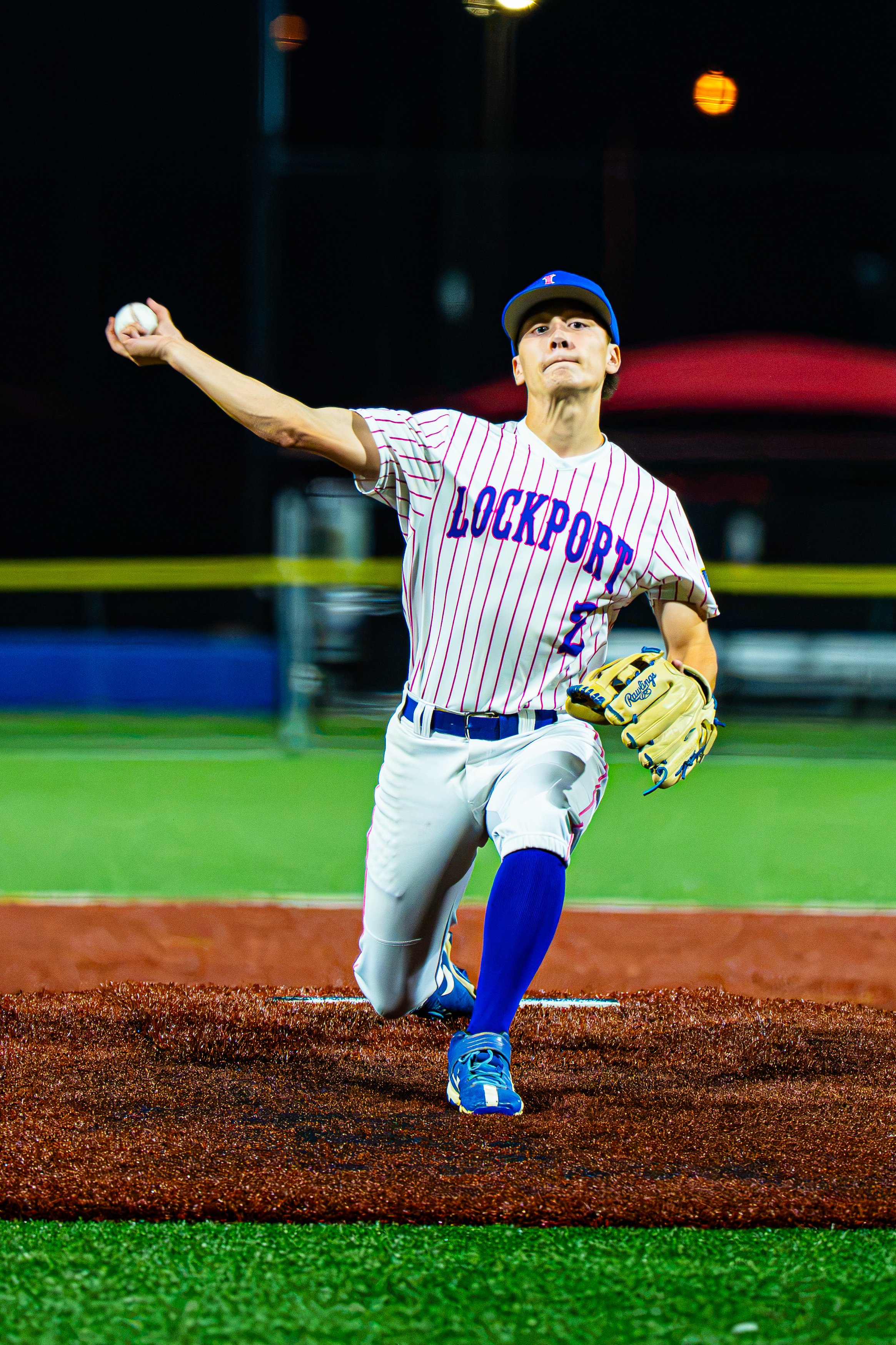A baseball player wearing a striped uniform with 'LOCKPORT' written on it, blue cap, and yellow glove, is pitching on a baseball field under stadium lights at night.