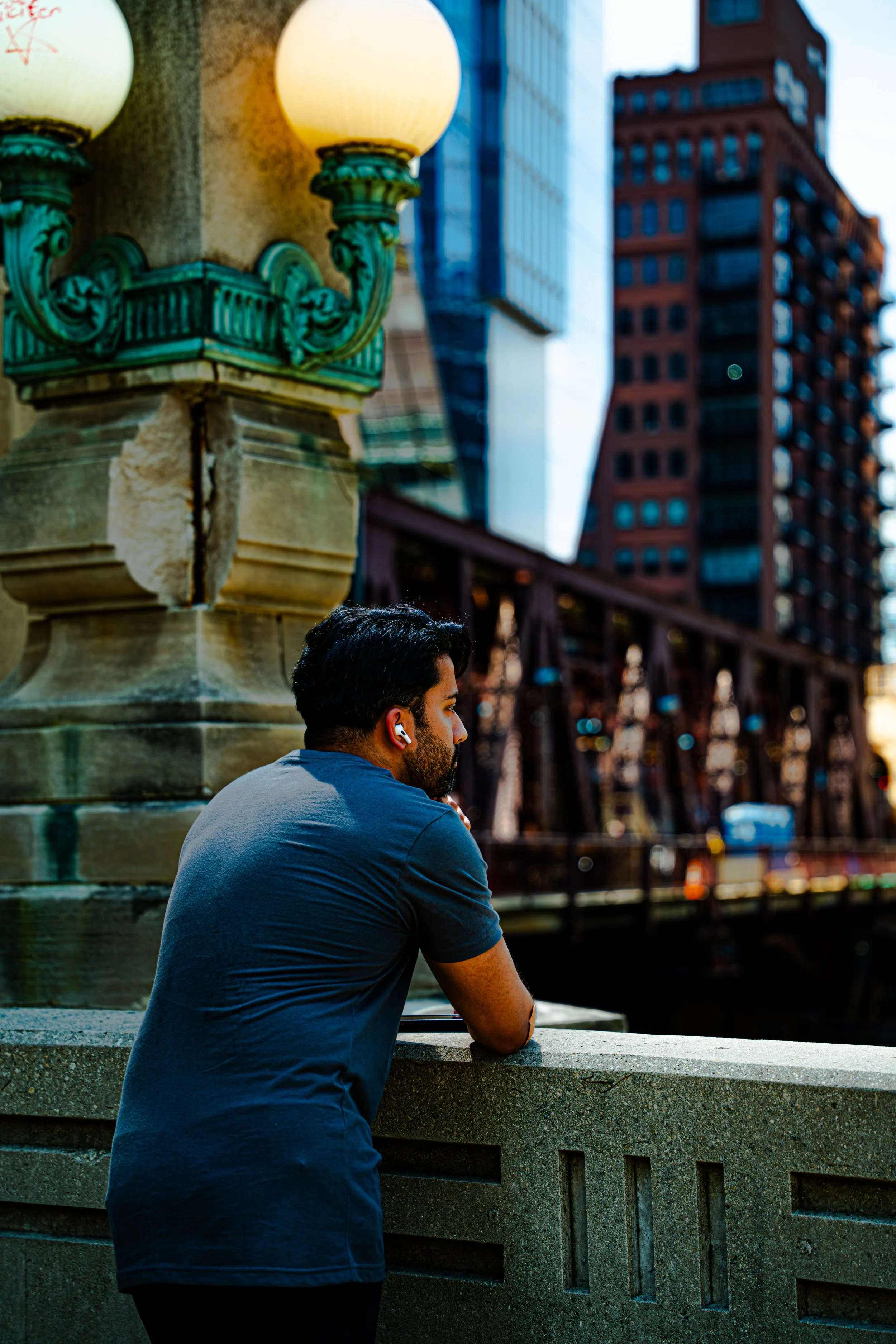 A man with dark hair, a beard, wearing a blue t-shirt, is sitting on a stone ledge with his arms resting on it, looking at a cityscape background with tall buildings including a red-brick skyscraper, during sunset. The man is listening to wireless ea