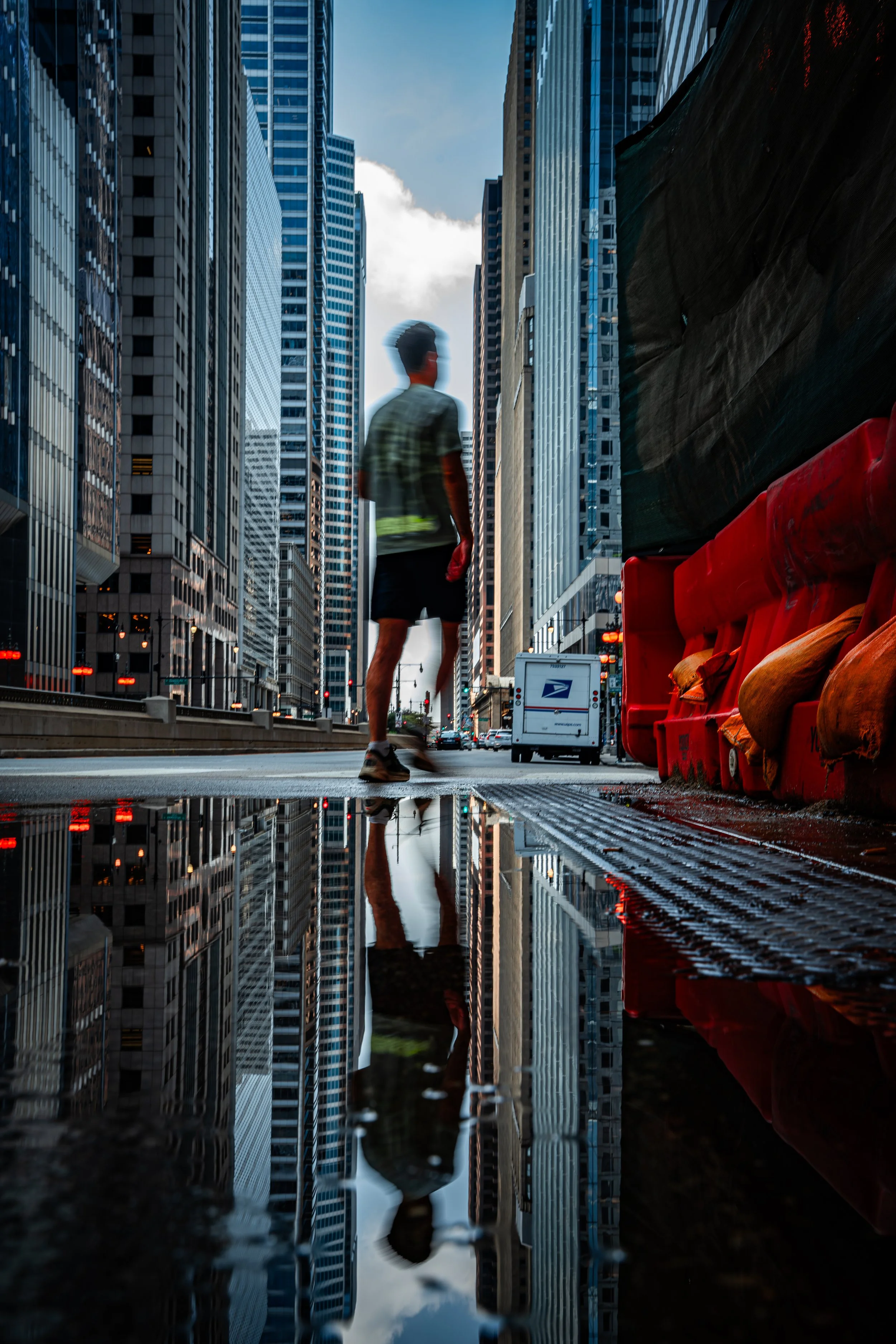 A person walking in a city street with tall glass buildings on either side. The reflection of the person and buildings can be seen on a wet surface on the ground. There are traffic lights and a postal service truck in the background.