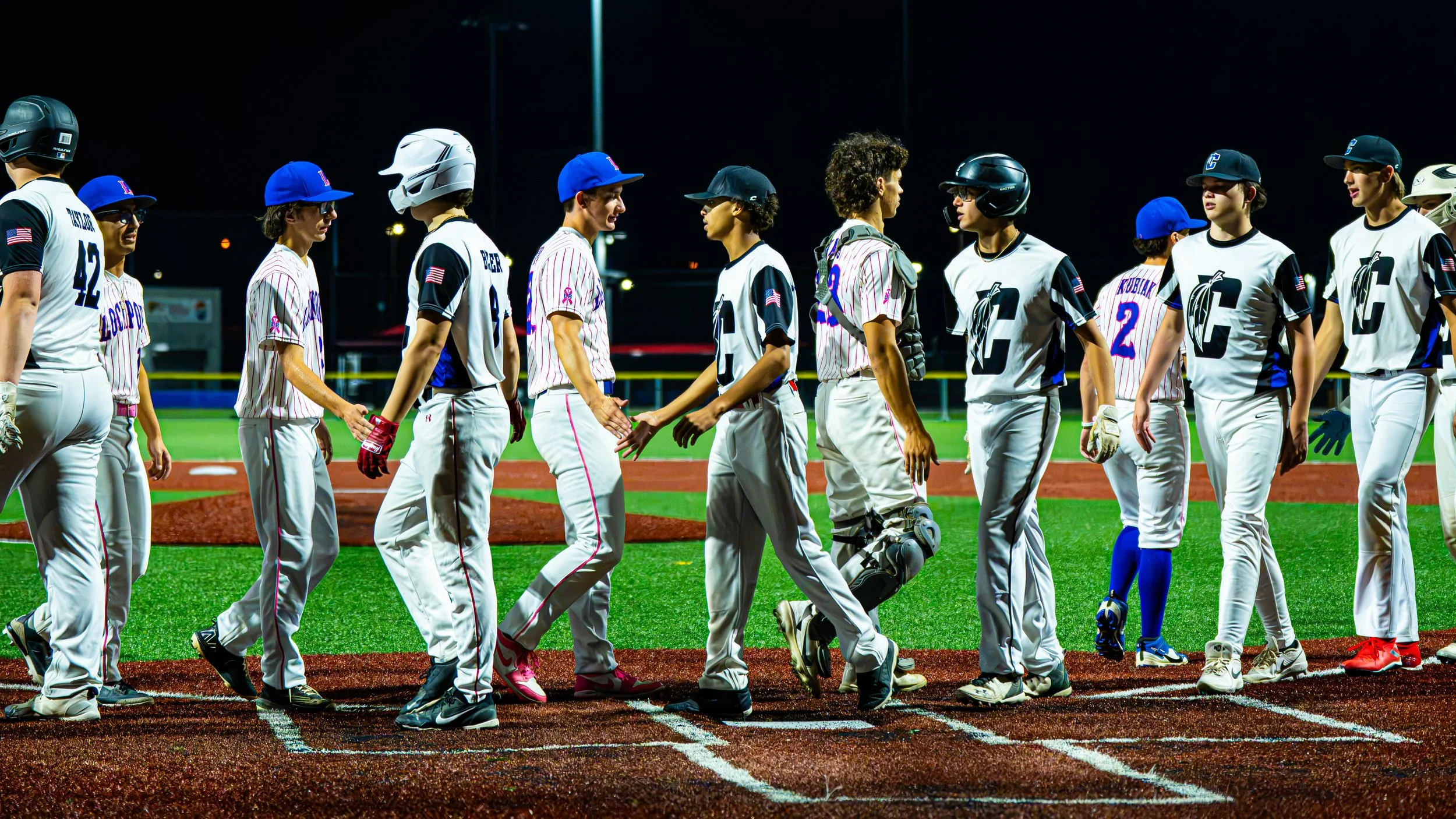 Nighttime scene of female and male baseball players standing on the field, shaking hands, some in uniforms and helmets, some with gloves, on a green artificial turf baseball field.