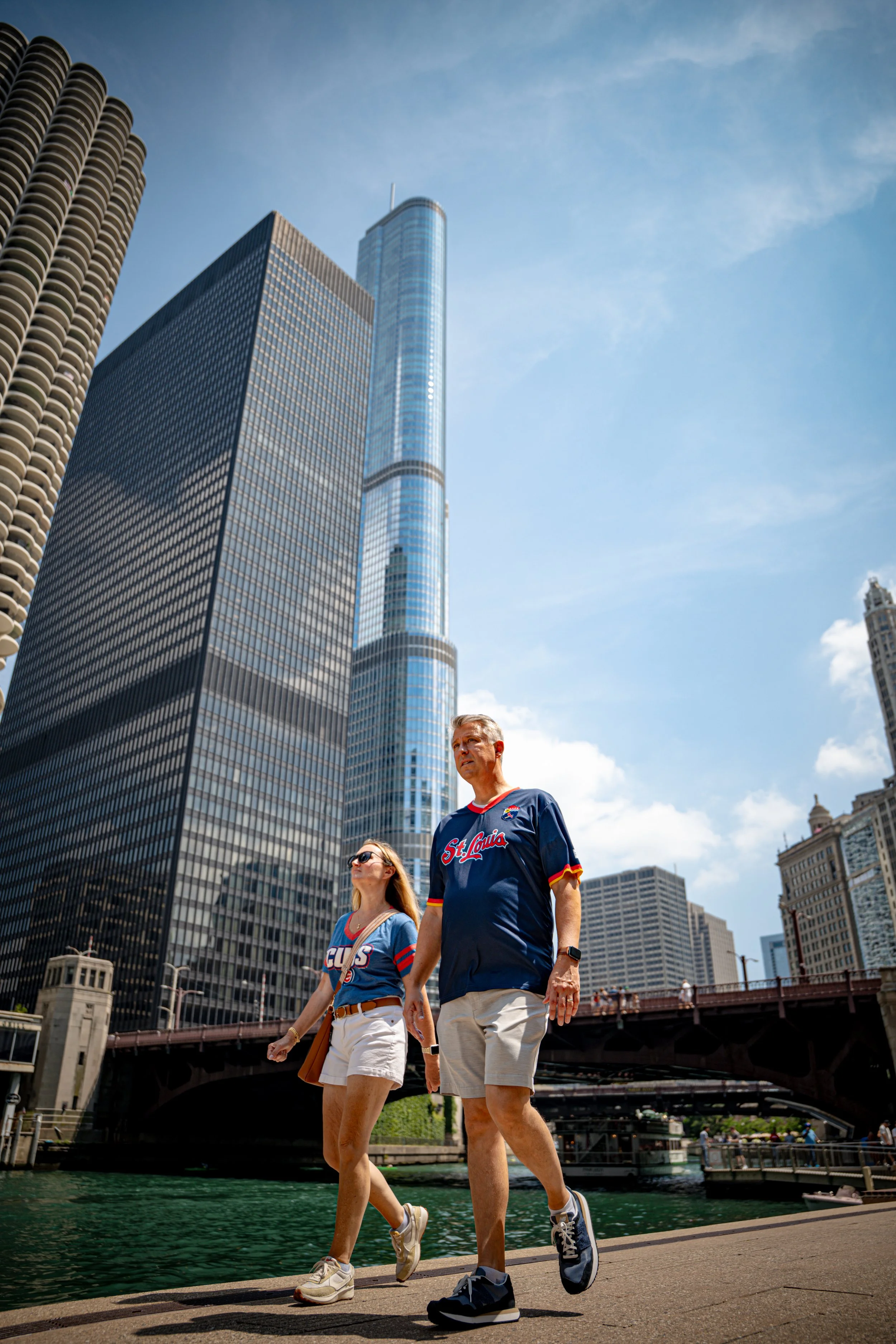 Two people walk along a riverfront in a city, with tall modern skyscrapers and a bridge in the background. One is a woman with long hair, wearing sunglasses, a blue jersey, and white shorts. The other is a man with short hair, wearing a blue sports j