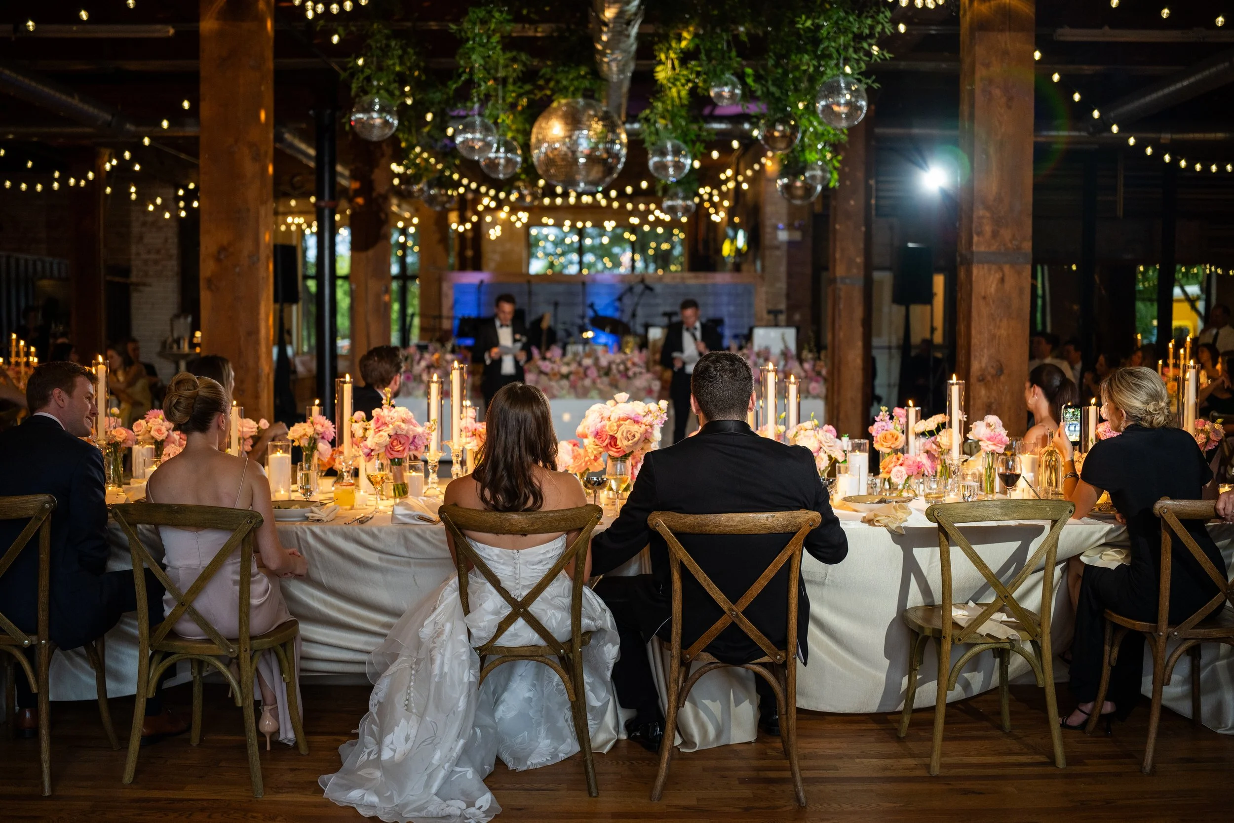 A wedding reception with guests seated at a long table decorated with pink flowers and candles, overlooking a band playing on a stage with hanging disco balls and string lights, in a rustic indoor venue with wooden beams.