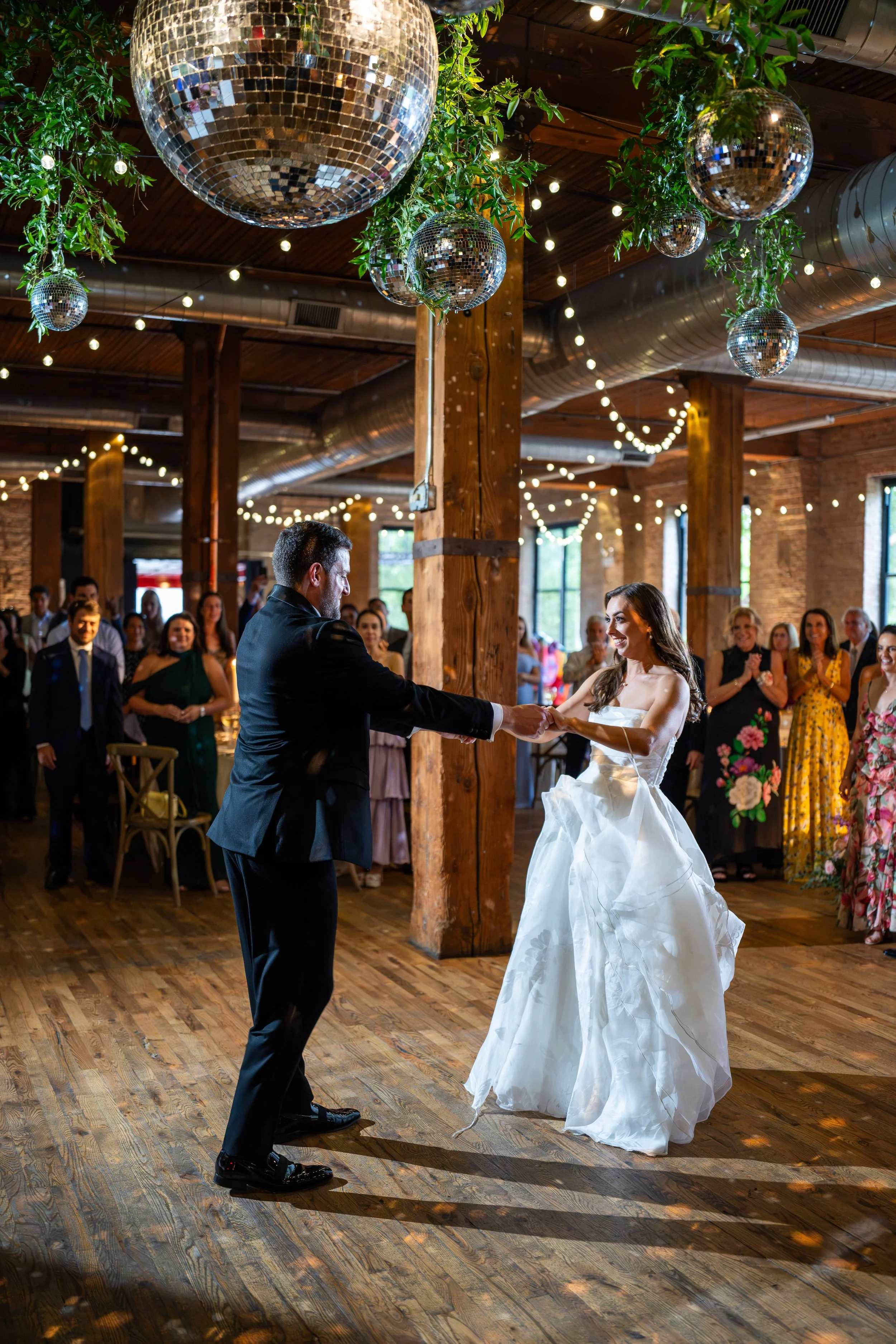 A bride and groom are dancing in a rustic indoor venue decorated with hanging disco balls and string lights, surrounded by guests watching and smiling.