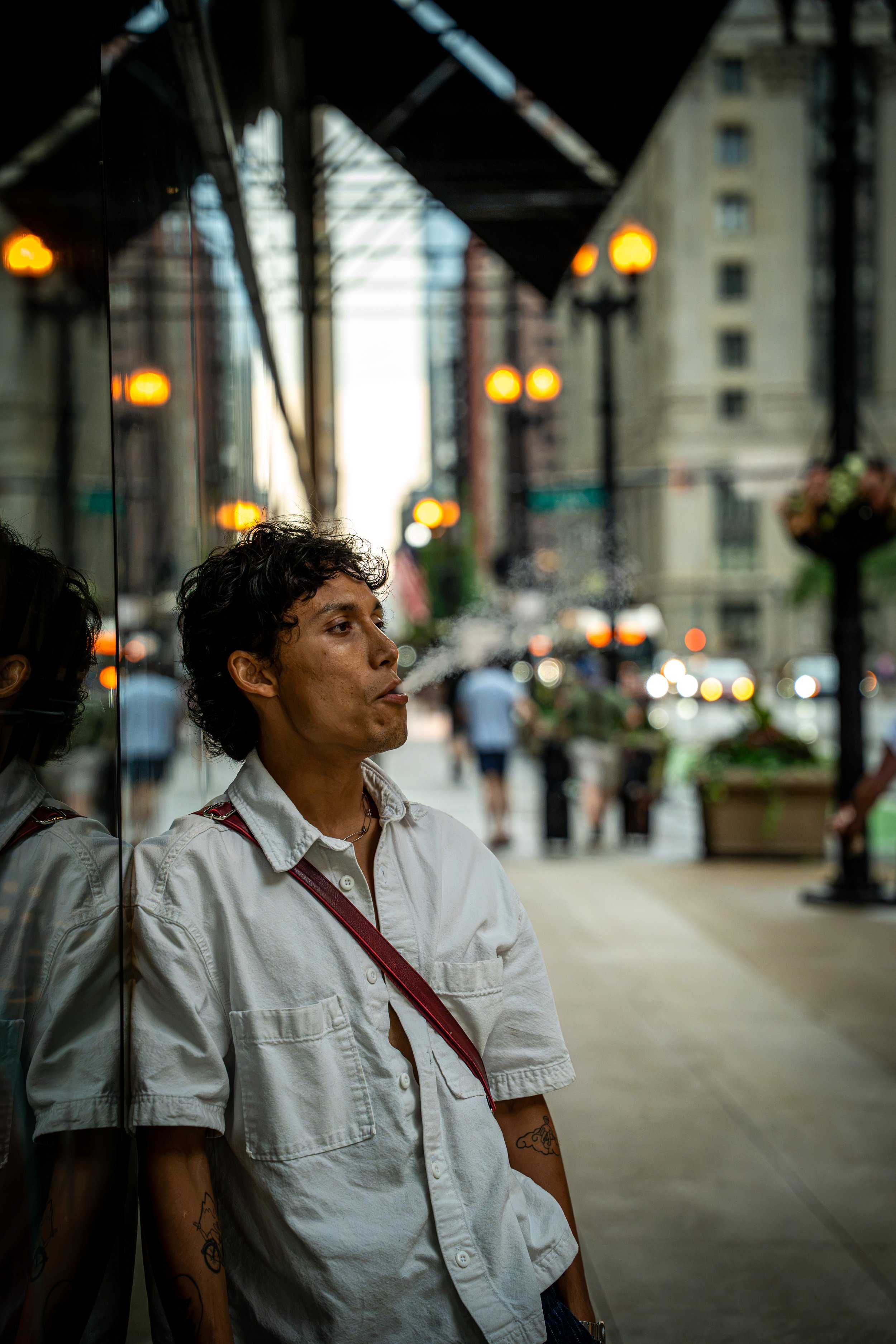 A young man with curly dark hair wearing a white short-sleeved button-up shirt and tattoos on his arms, leaning against a reflective surface on a busy city street.