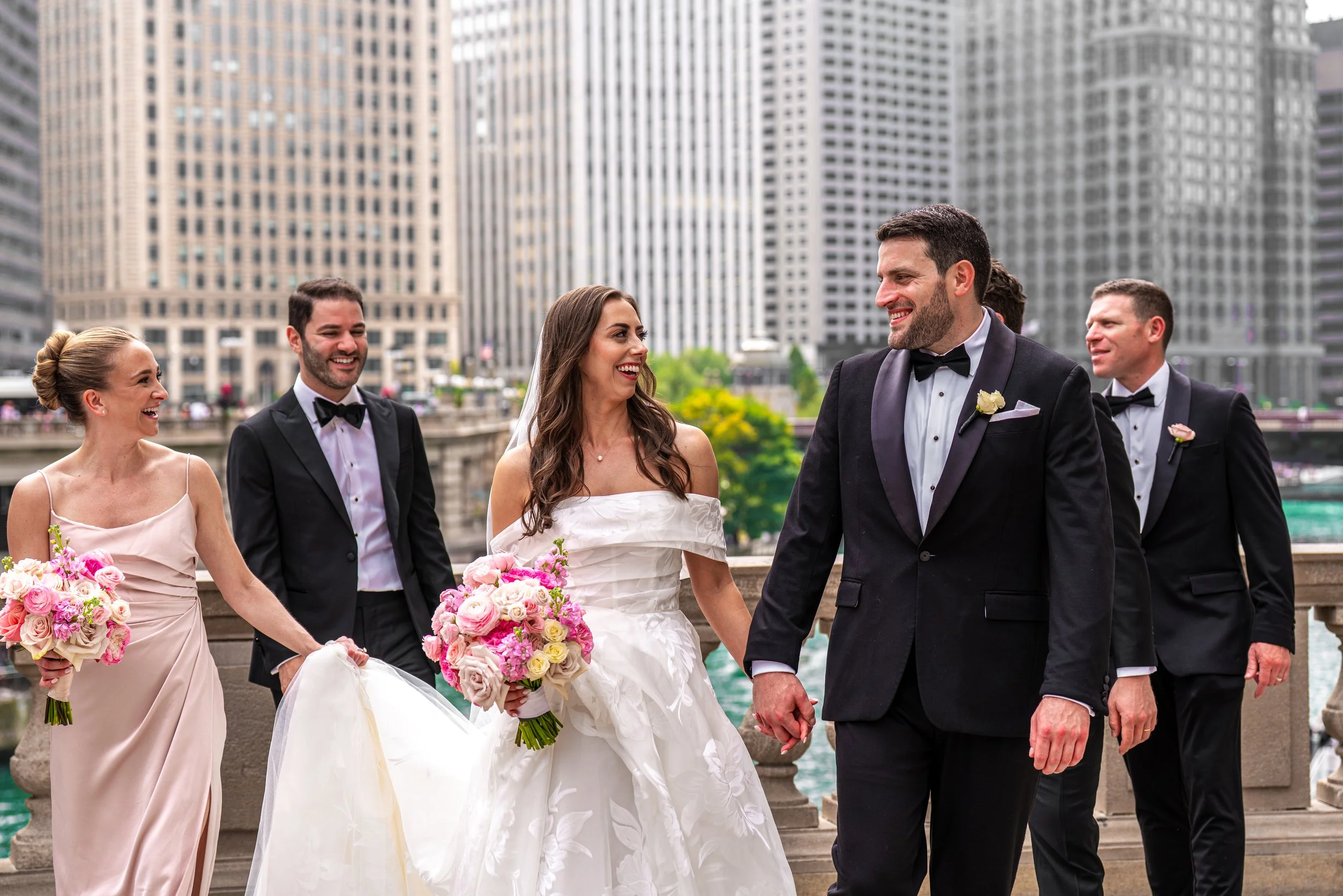 A group of smiling people dressed in formal wedding attire, including a bride in a white gown holding a bouquet of pink and cream roses, walking outdoors in an urban setting with tall buildings in the background.