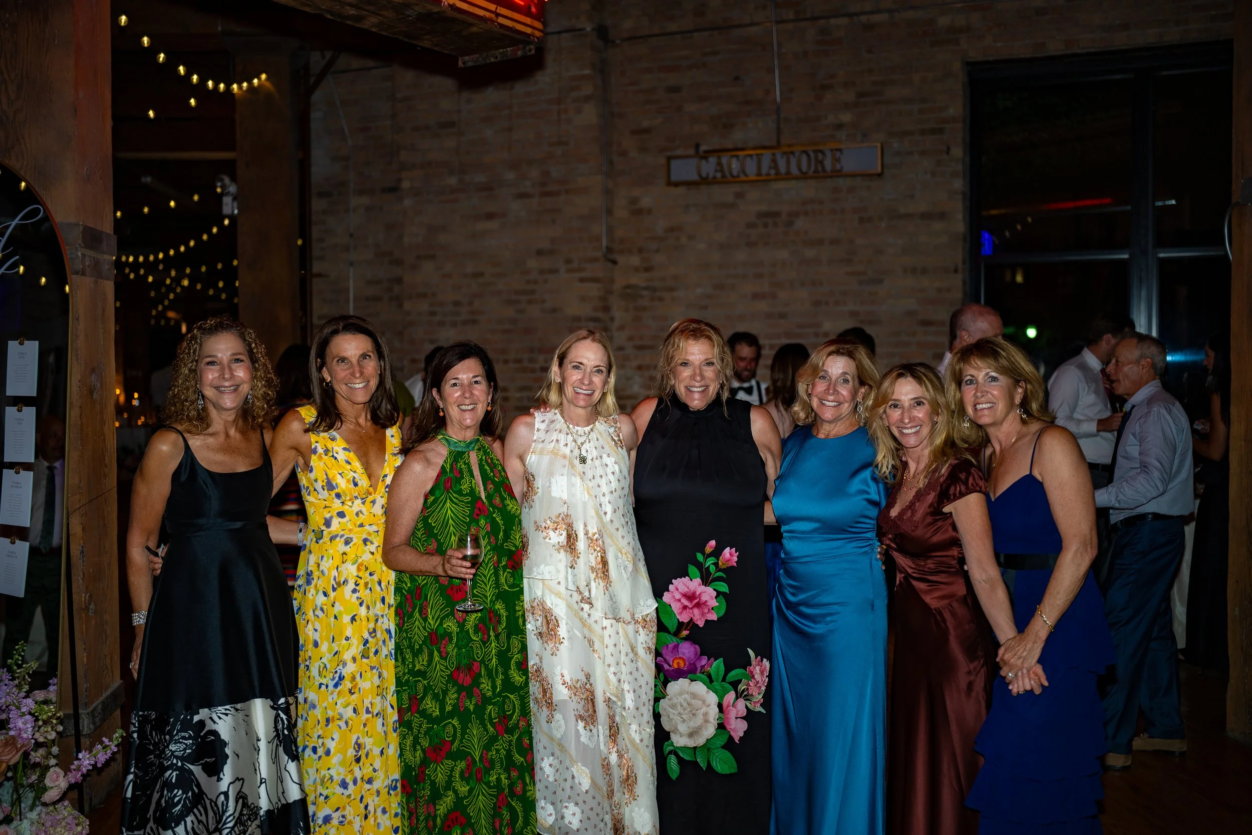 Group of women dressed in colorful formal attire, smiling and posing together at a social event in a brick-walled venue with string lights.