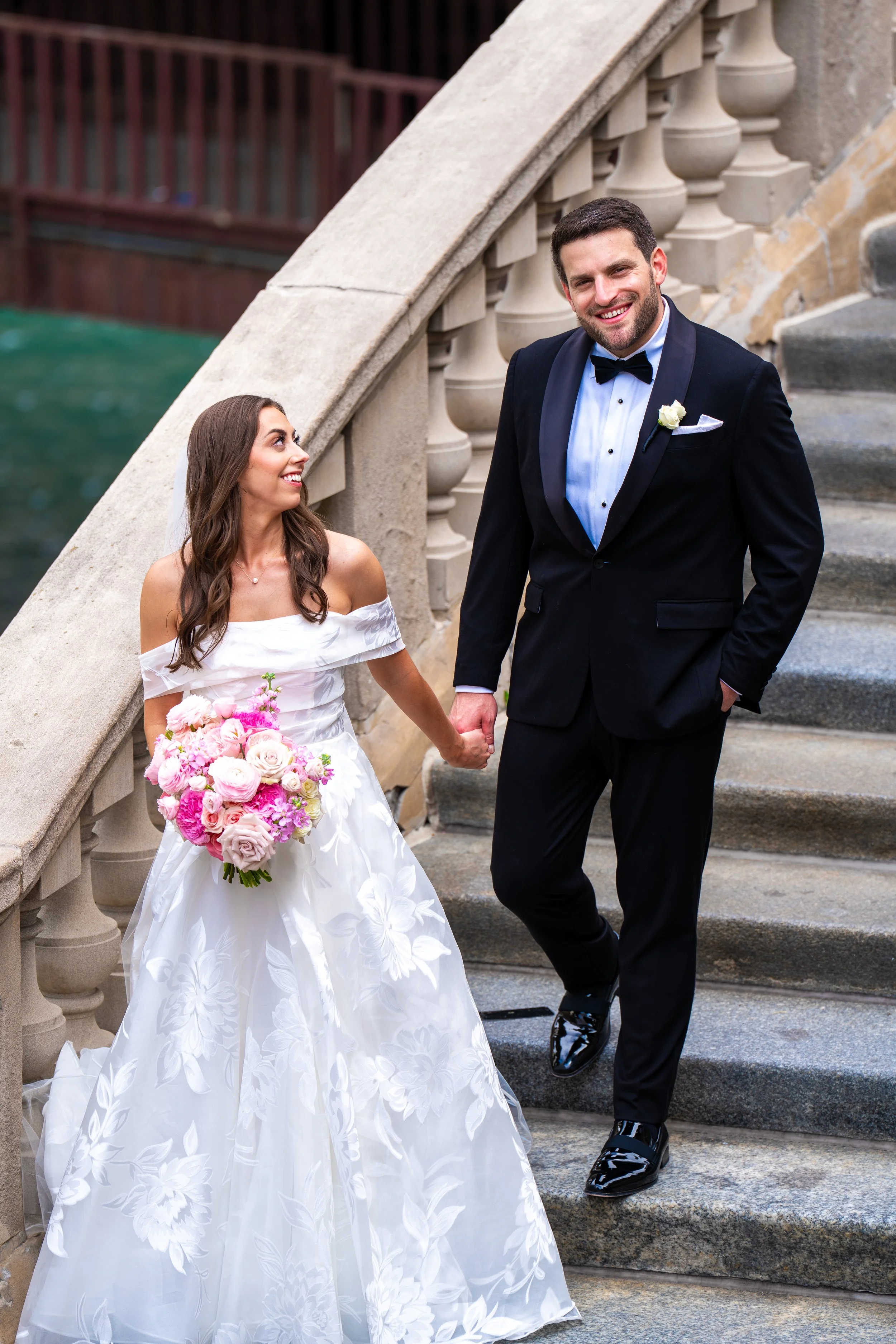 A bride and groom walking hand in hand down a stone staircase outdoors. The bride wears a white wedding dress with floral patterns and holds a bouquet of pink and white flowers. The groom wears a black tuxedo with a bow tie and a white boutonniere, smiling at the camera.