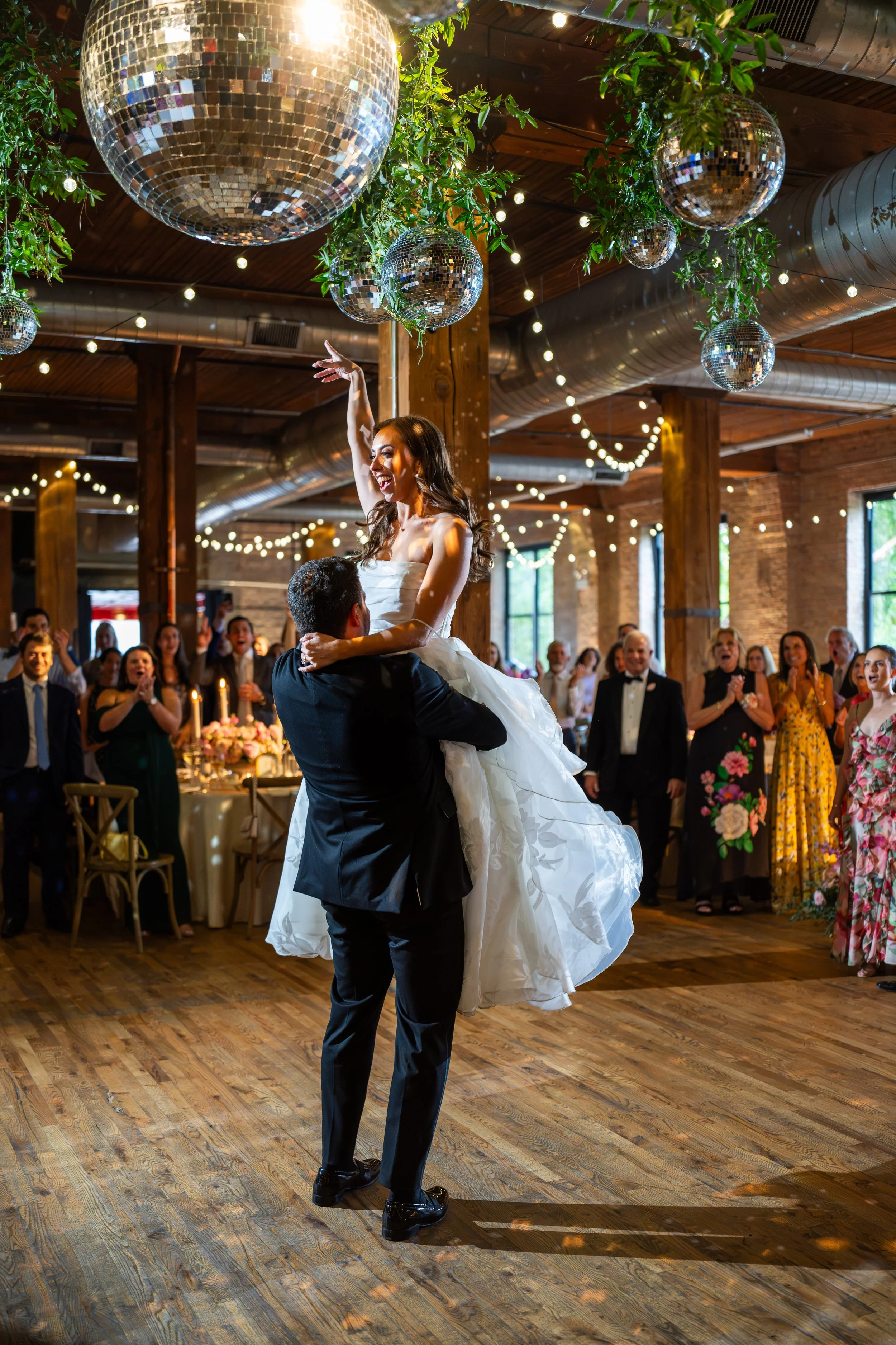 A couple dances at a wedding reception, with the woman lifted in the air and smiling. The venue has wooden floors, large dance floor, and disco balls hanging from the ceiling. Guests are clapping and celebrating in the background.