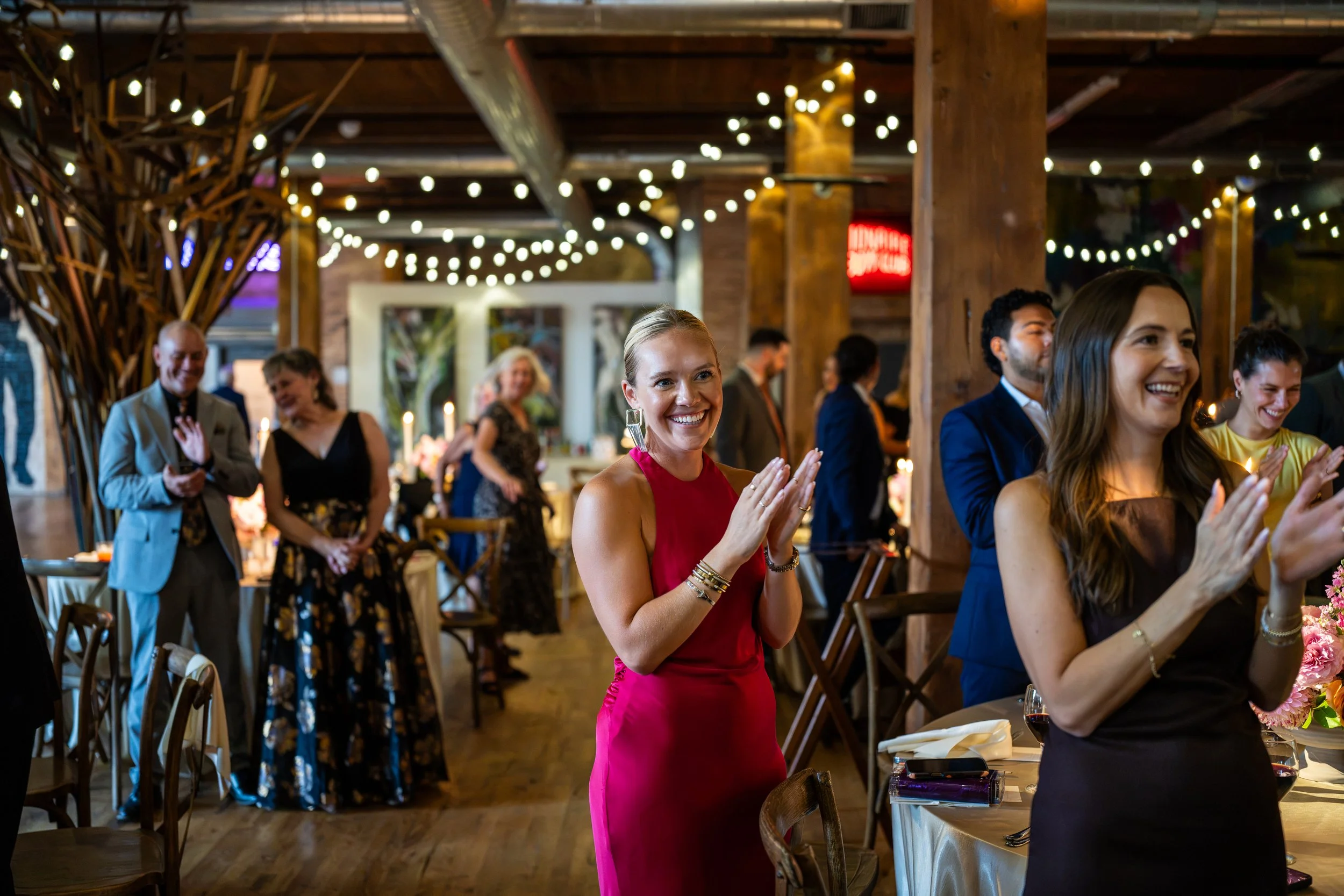 A group of people at a celebration or party inside a rustic venue with string lights, with some people clapping and smiling.