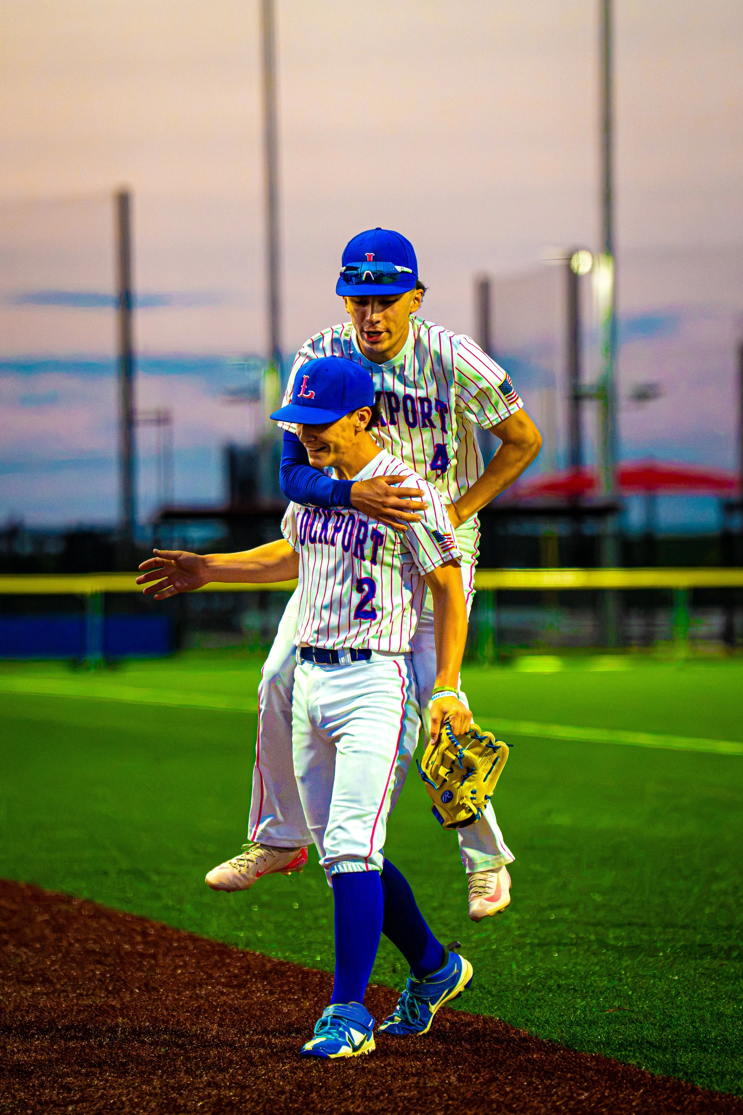 Two young baseball players in white and blue uniforms celebrating on the field during sunset, with one wearing a blue cap and holding a glove, the other on a mound of dirt, with a baseball field and sky in the background.