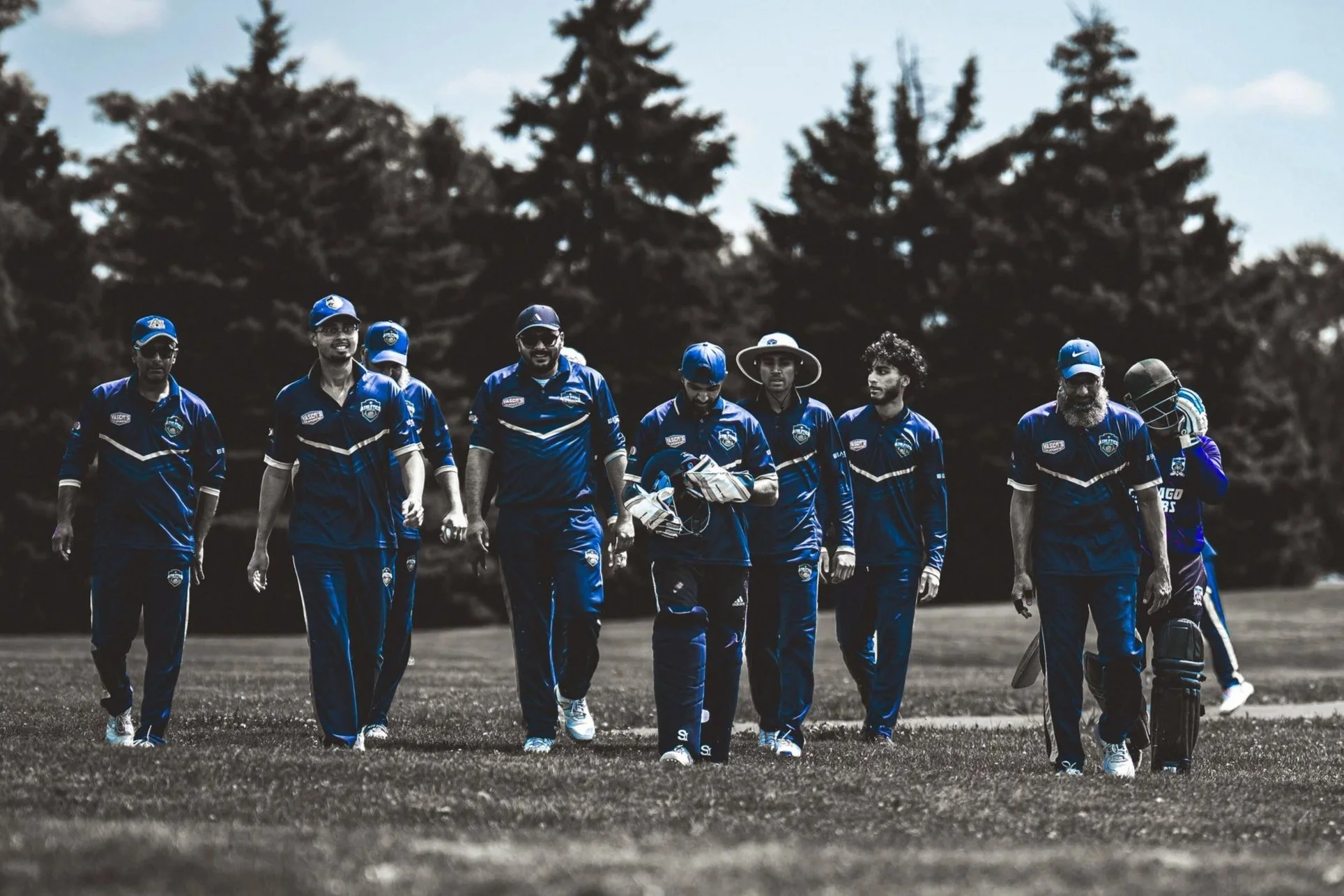 Cricket team walking on field wearing blue uniforms with caps and helmets, surrounded by trees in the background.