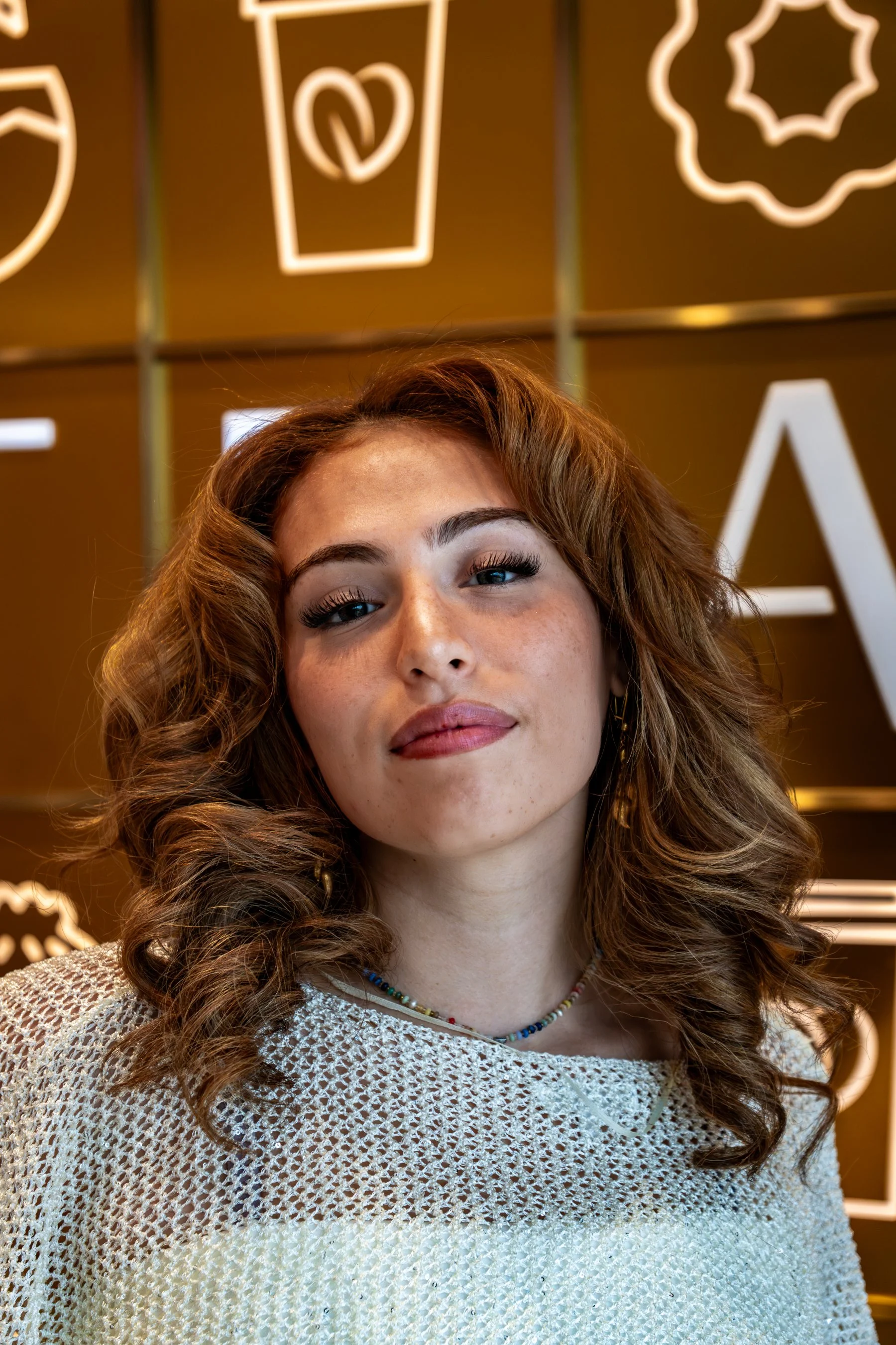 Close-up image of a young woman with curly brown hair, wearing a white crochet top and a colorful beaded necklace, standing in front of a brown background with white neon signs.