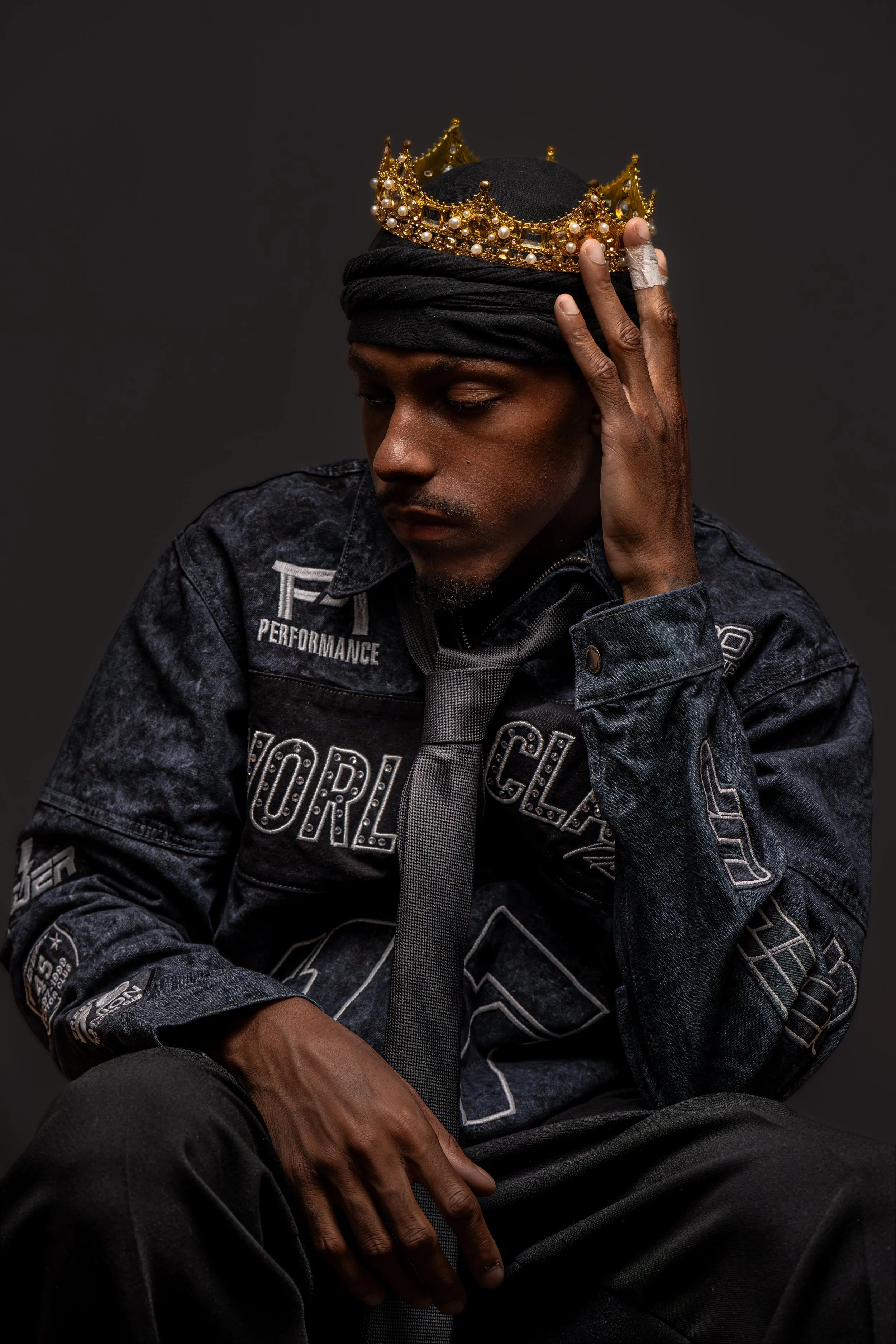 A man with dark skin wearing a black headwrap and a denim jacket with patches, sitting with eyes closed and holding a gold crown adorned with pearls on his head, against a dark background.